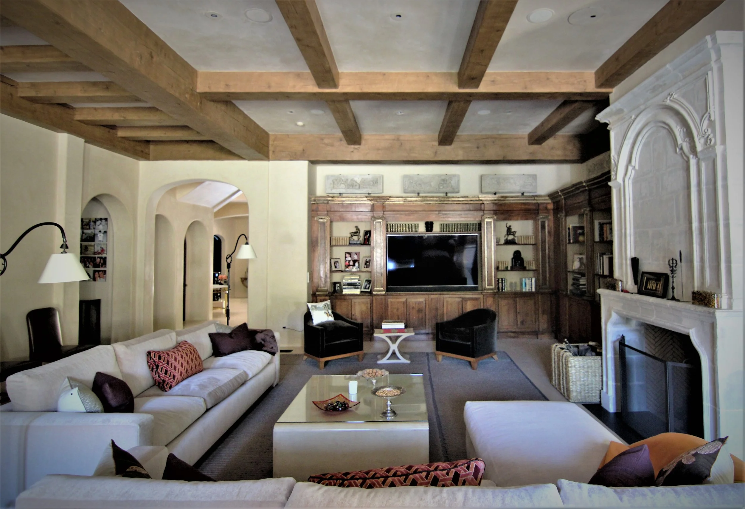 Living room with cream-colored sofas, a fireplace, built-in wood shelves, a large TV, and a wooden ceiling with exposed beams.