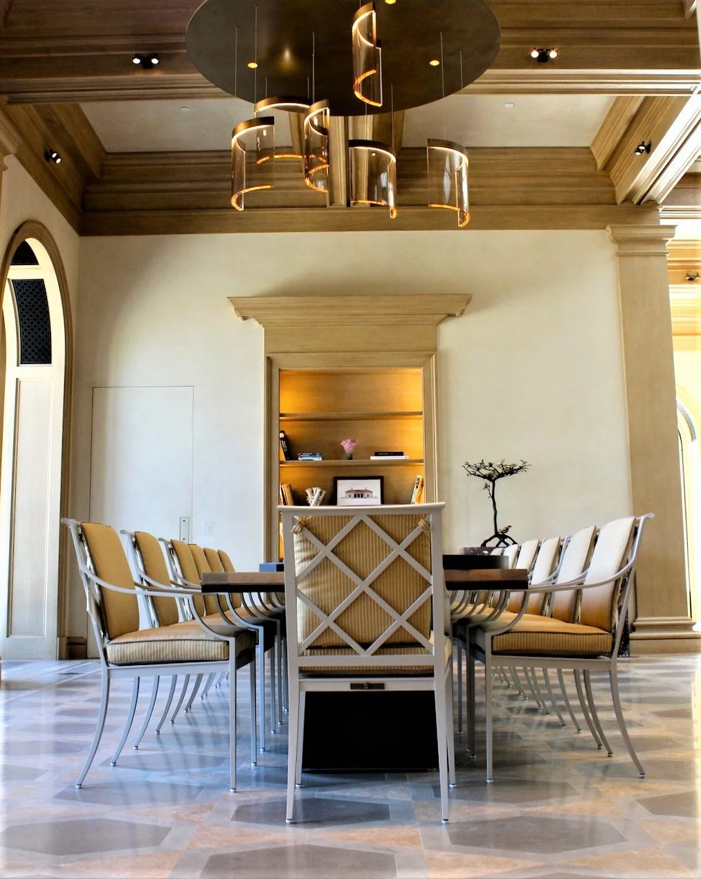 Elegant dining room with a large rectangular table, surrounded by beige cushioned chairs with metal frames, and a decorative wooden bookshelf in the background. The ceiling has a modern chandelier with gold accents, and there is a small bonsai tree o