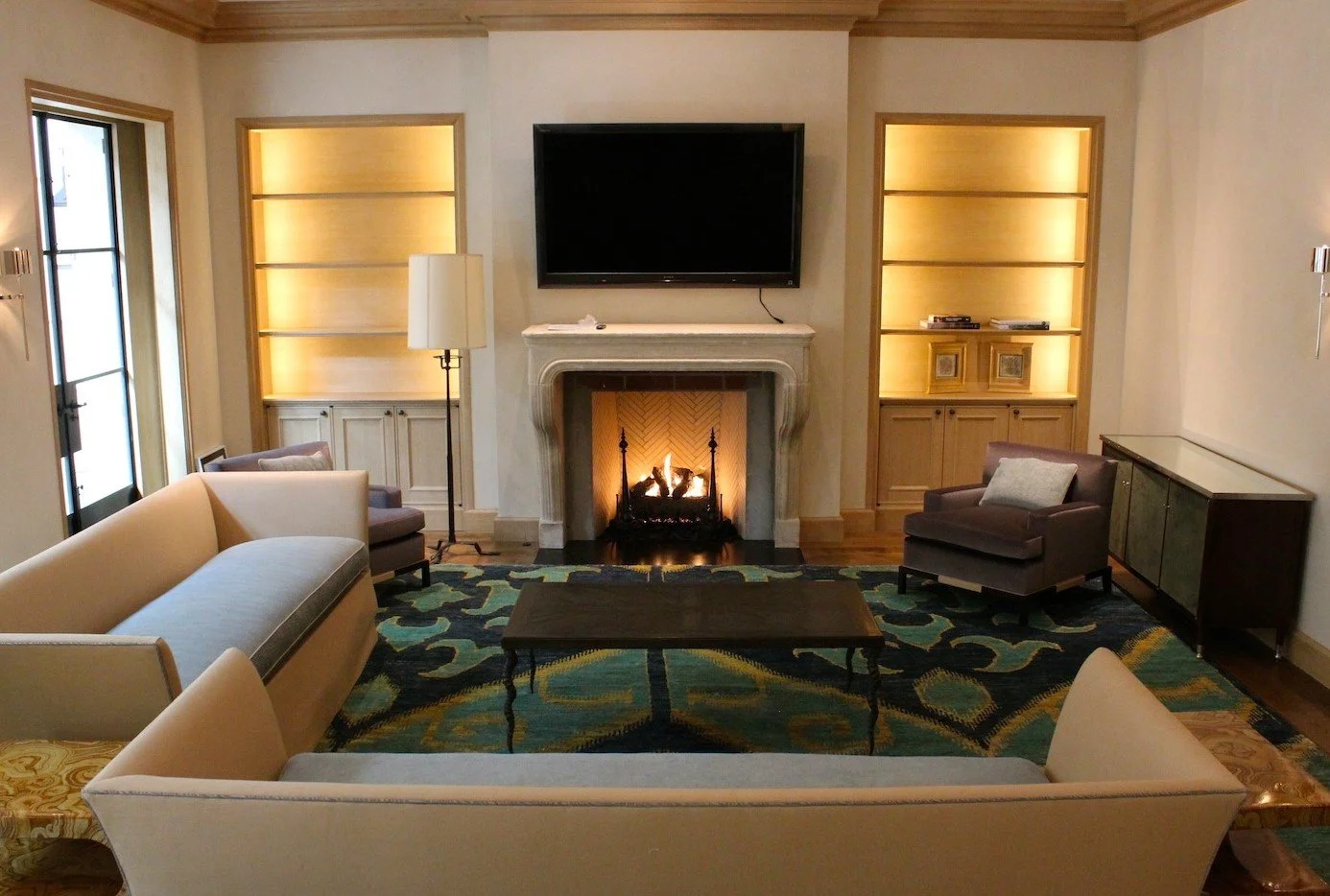 Living room with beige and gray furniture, a fireplace, a wall-mounted TV, built-in shelves with lighting, a patterned rug, and windows with natural light.