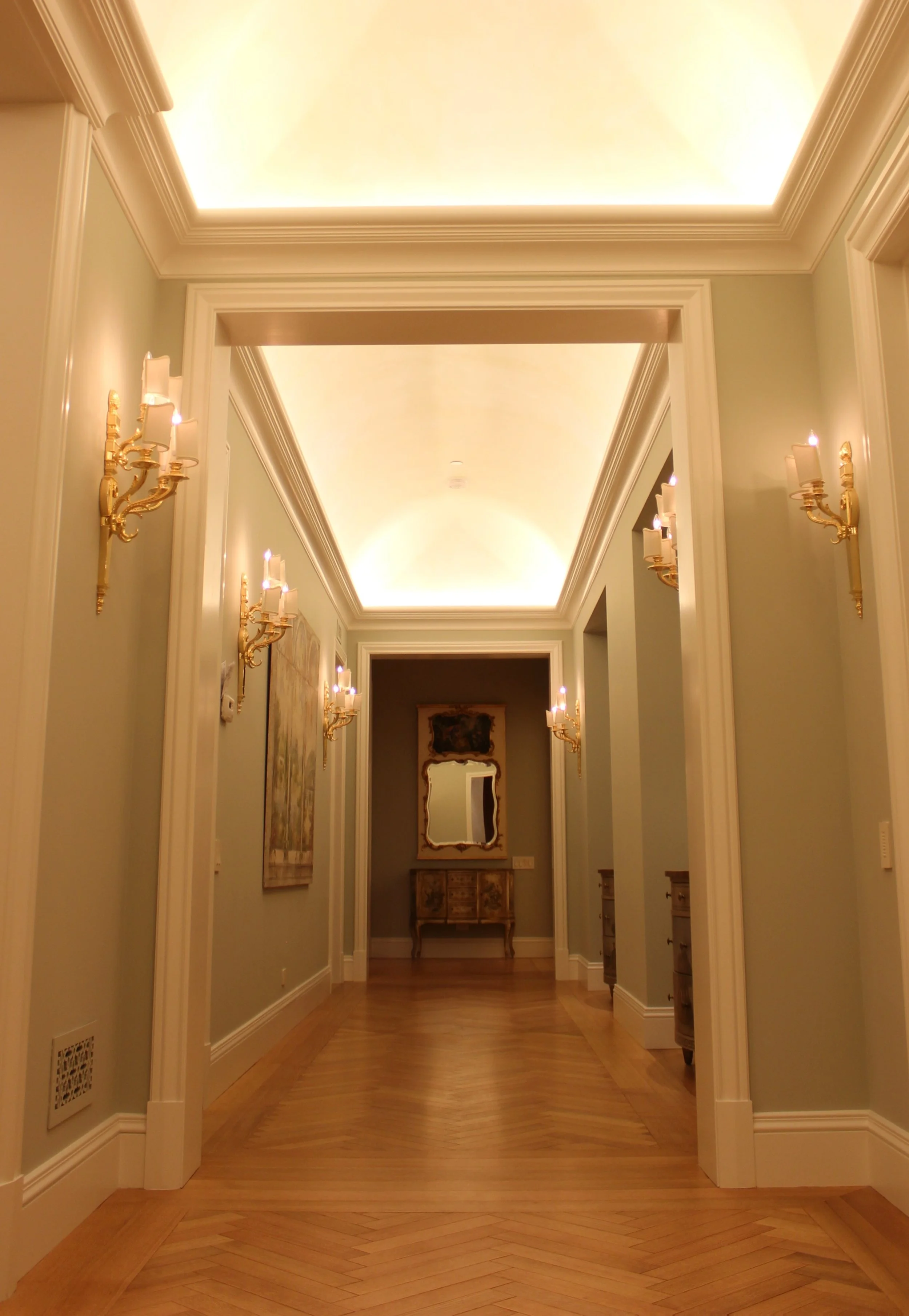 Elegant hallway with soft yellow lighting and gold wall sconces, classical artwork, and a mirror, with polished wooden flooring and white trim.