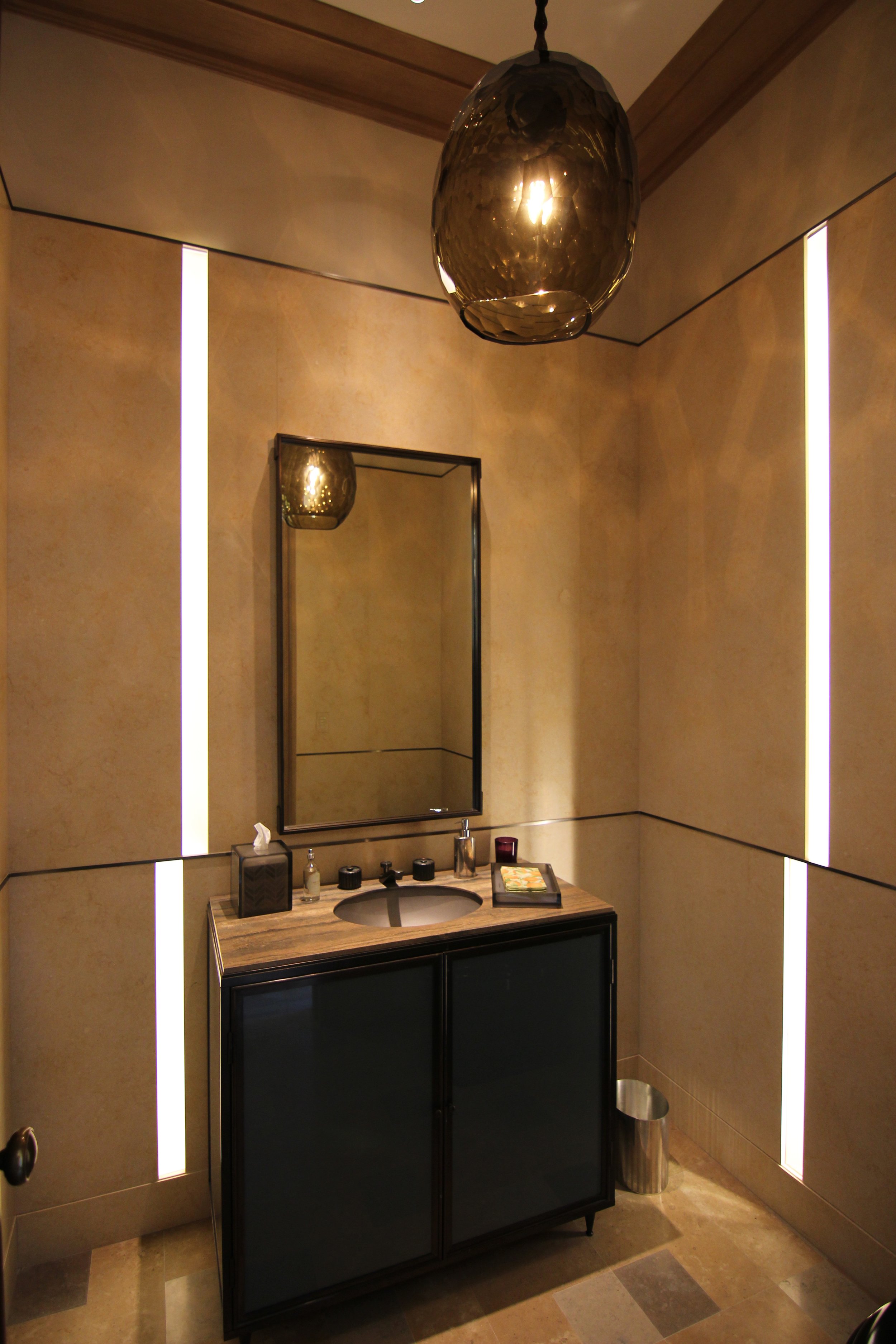 A modern bathroom with beige walls, vertical LED light strips, a rectangular mirror, a black vanity cabinet with a wood countertop, a sink, soap dispensers, a tissue box, a lit candle, and a small trash can.