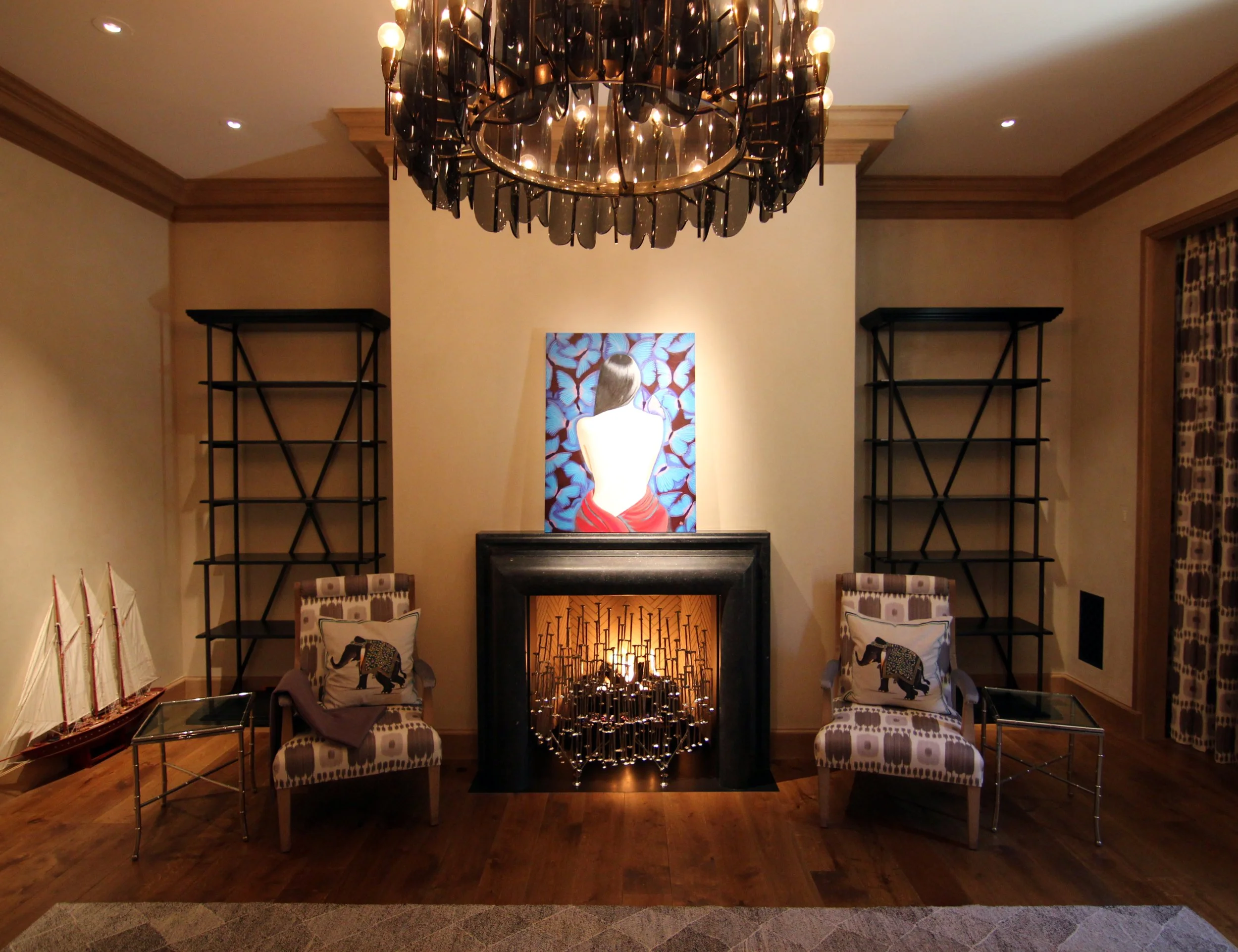 A living room with a fireplace, two patterned chairs with elephant pillows, glass side tables, and a nautical ship model. There are black shelves on either side of the fireplace, a colorful painting of a woman with her back turned above the fireplace