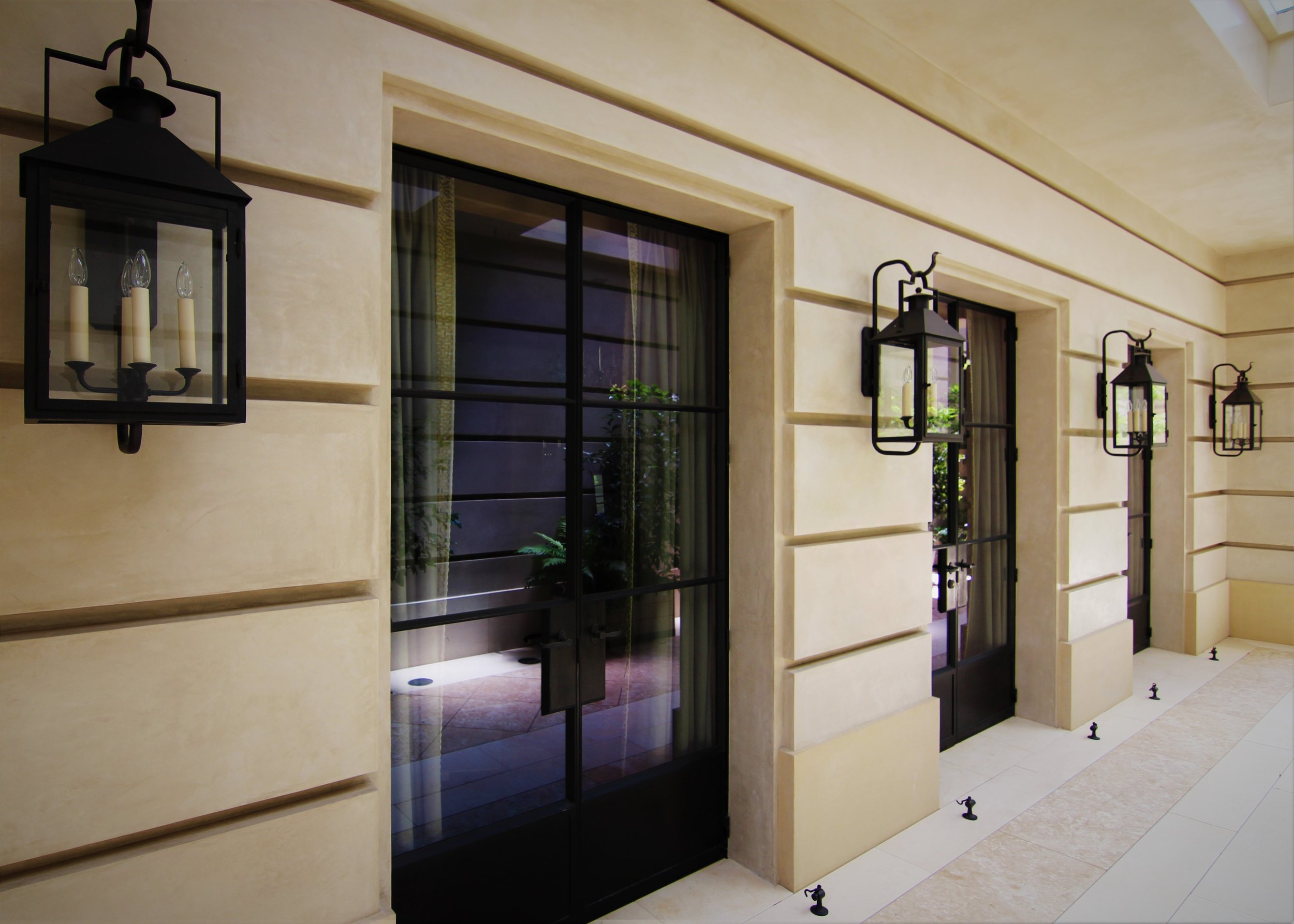 Exterior hallway with three black lantern-style wall sconces on a beige wall, glass doors, and small black decorative anchors at the base of the wall.
