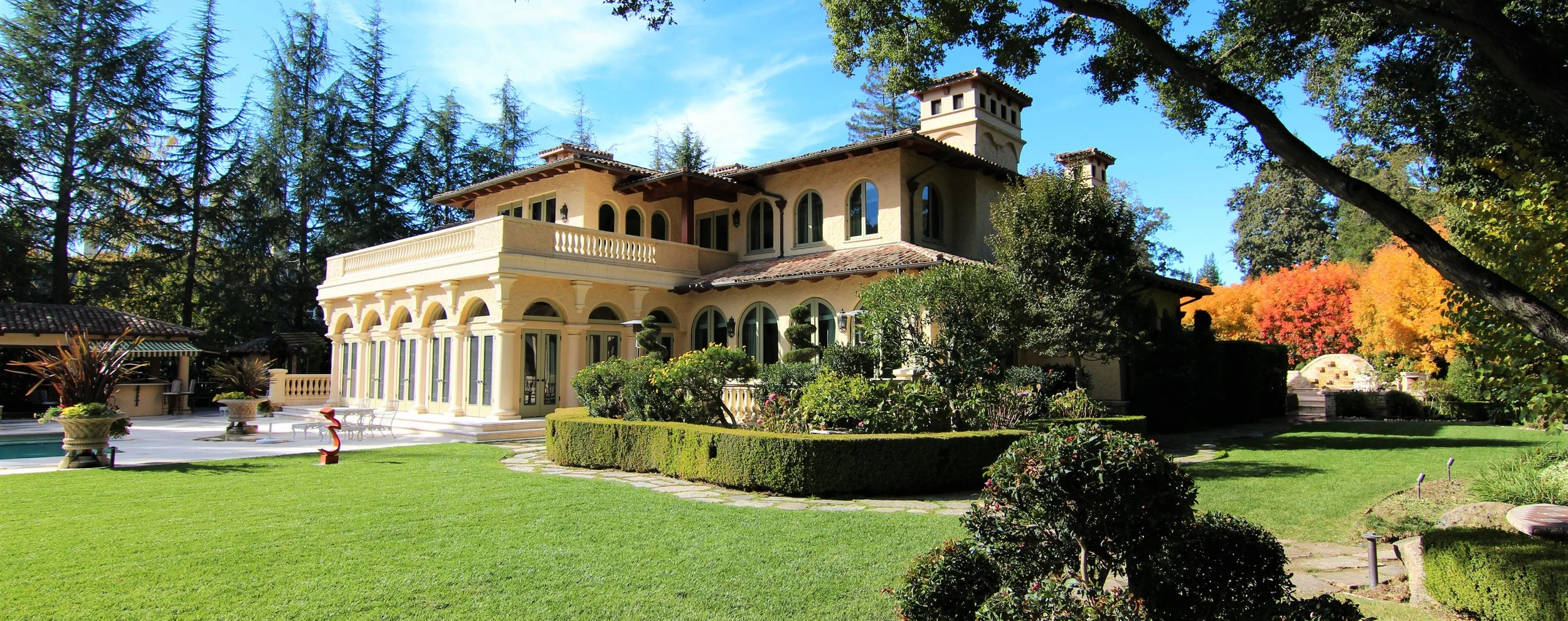 A large mansion with Mediterranean-style architecture, surrounded by a manicured lawn, colorful trees, and landscaped bushes, under a blue sky.