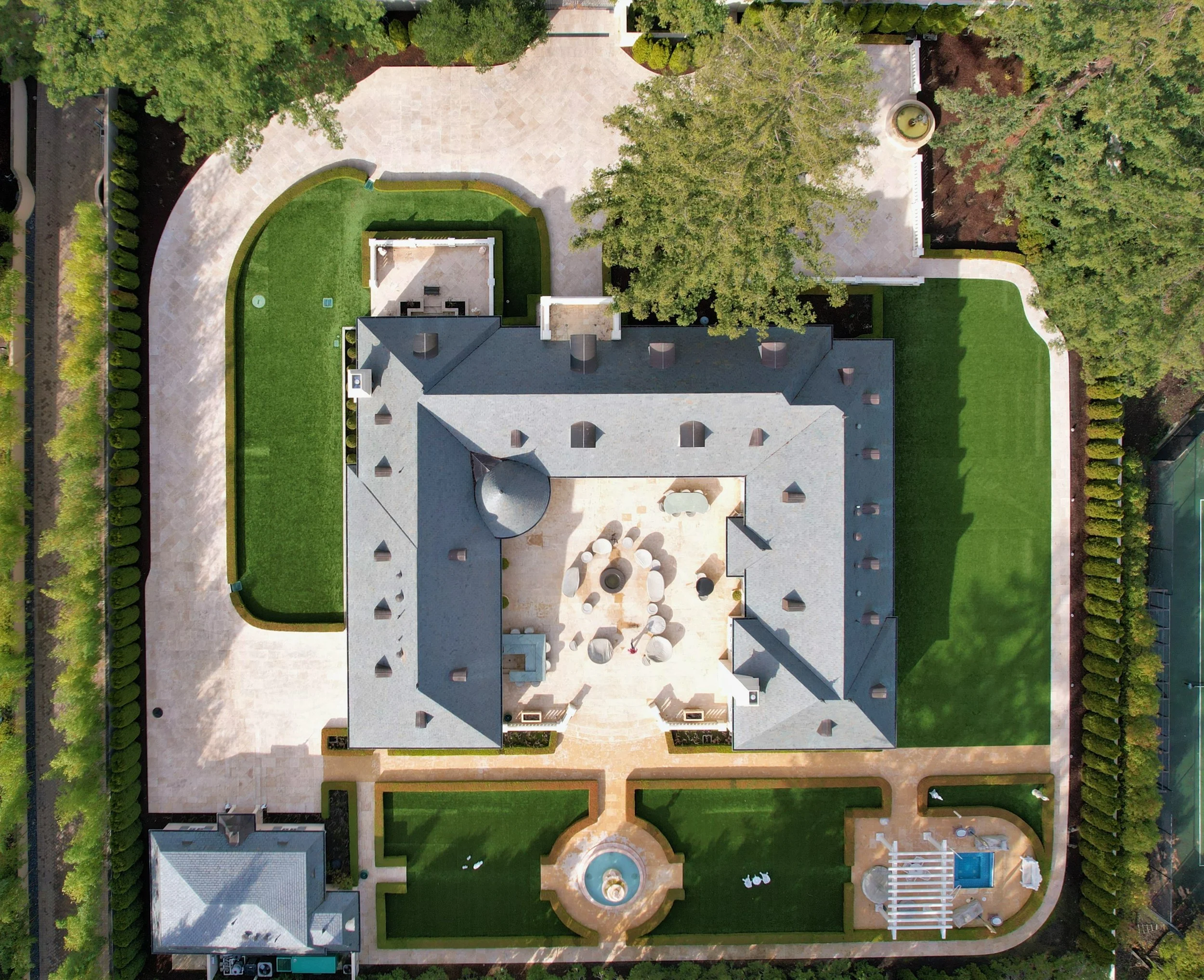 Aerial view of a large estate with a gray roof, surrounded by a well-maintained lawn, paved walkways, and tall trees. The estate features a central courtyard with outdoor seating, a fountain at the front, and a swimming pool with a pergola at the bac