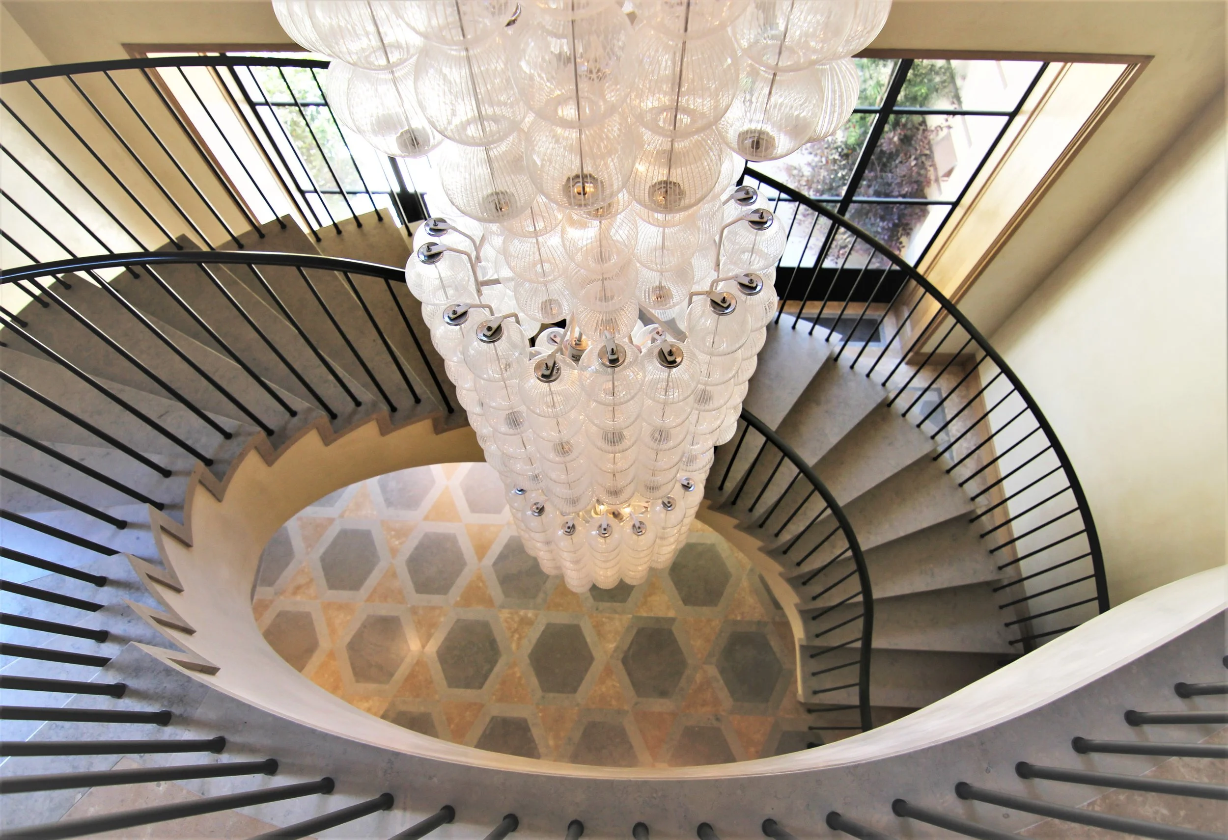 Inner view of a spiral staircase with black railings and a large chandelier made of multiple white glass globes hanging from the ceiling, with a window showing trees outside.