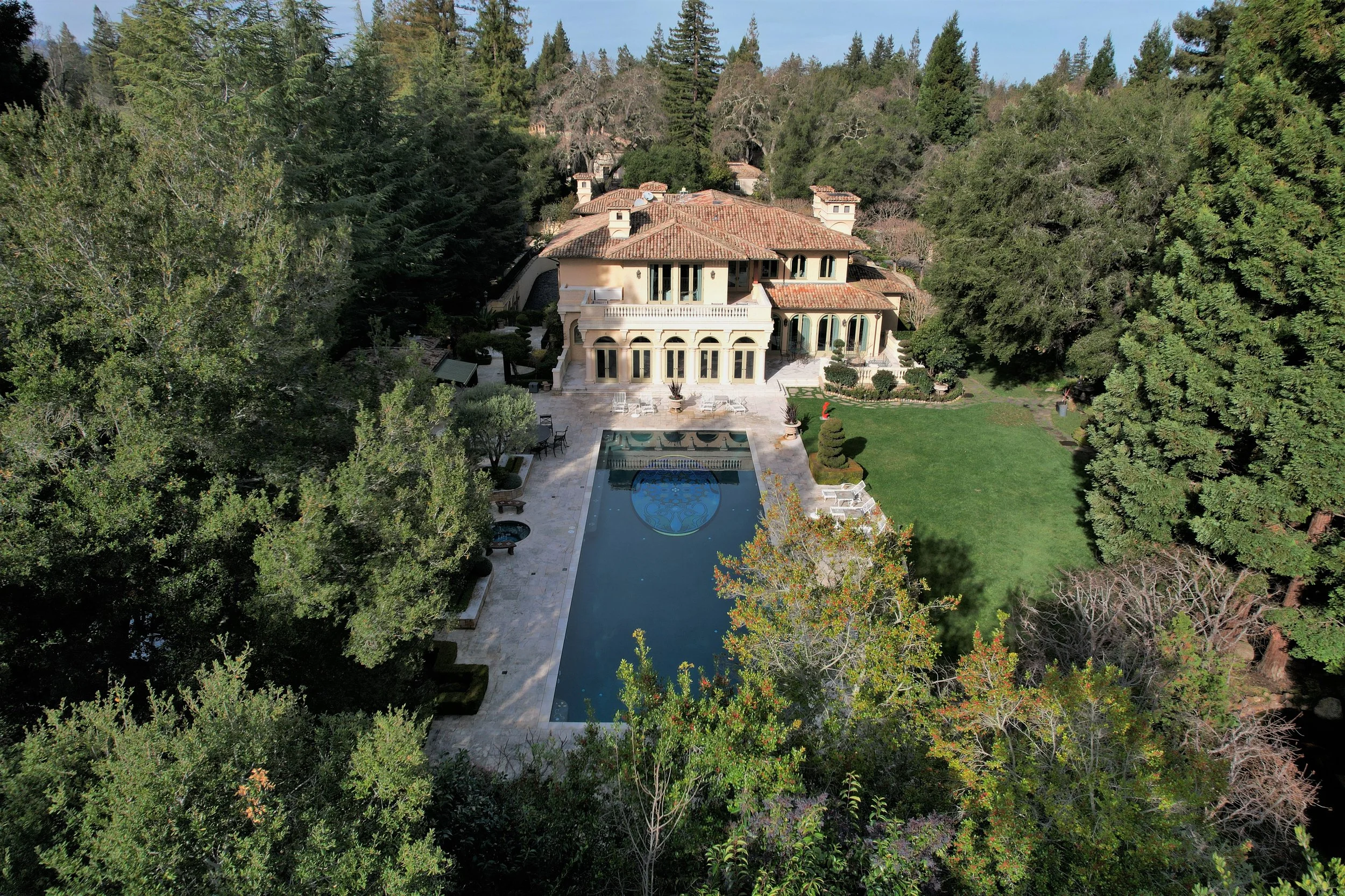 A large luxurious house with multiple floors, a swimming pool in the backyard, and surrounded by tall trees and a well-manicured lawn.