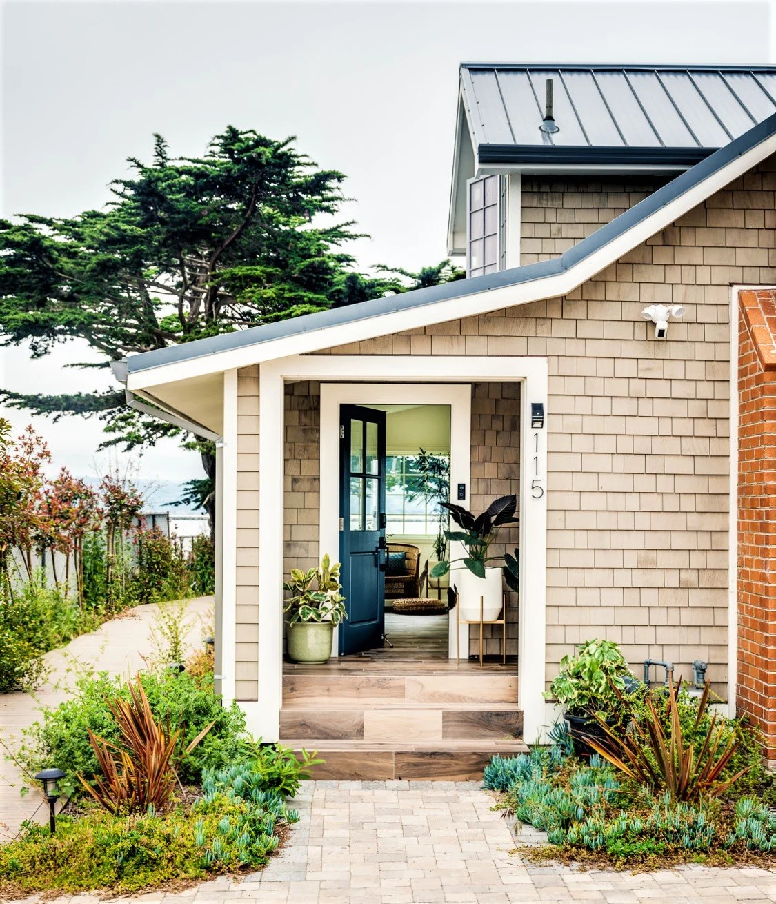 Front view of a modern house with a porch, potted plants, a blue door, and a garden path, featuring a beige shingle exterior and a gabled metal roof.
