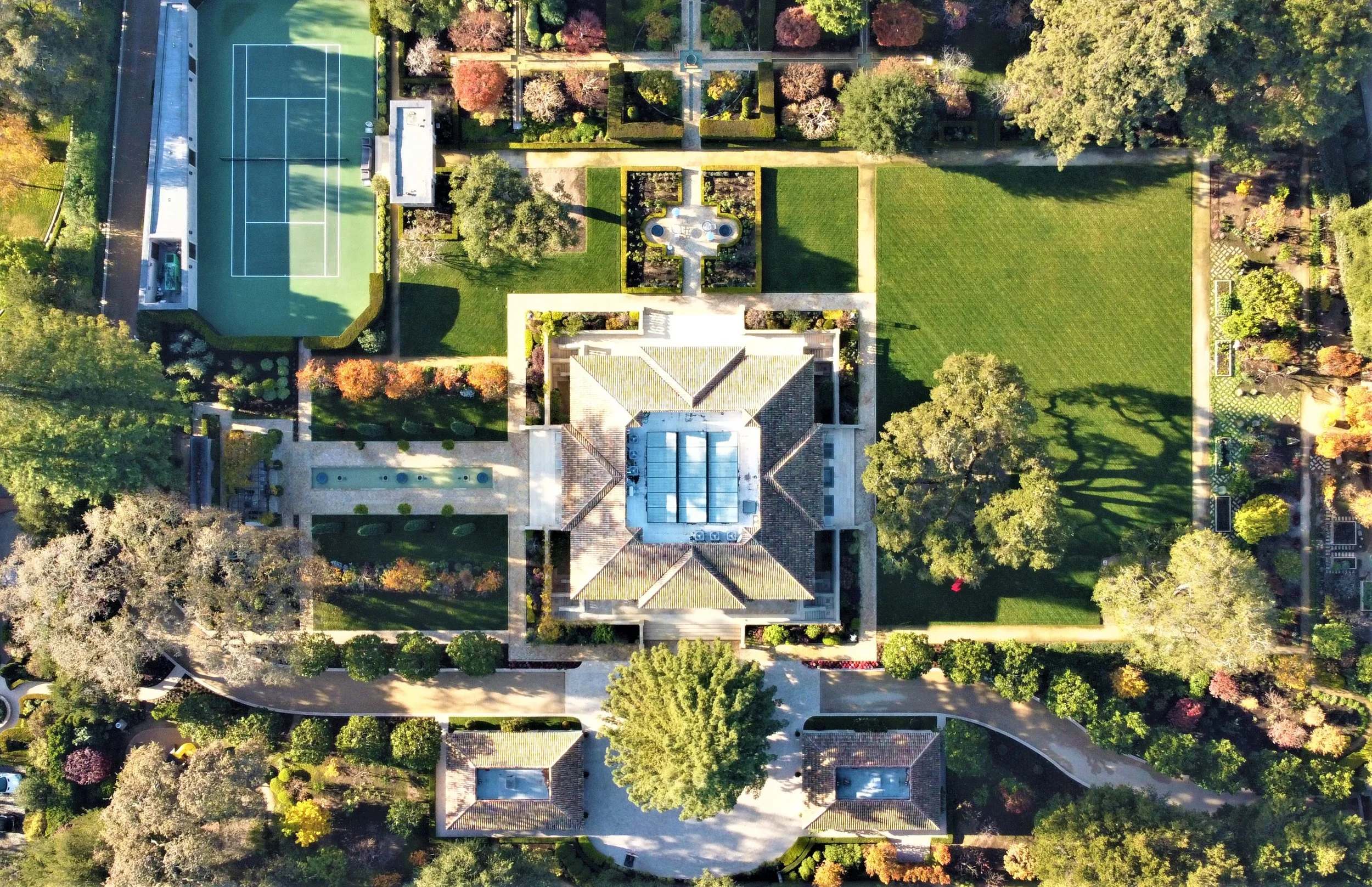 An aerial view of a large estate with a central house, tennis court, swimming pool, garden, and expansive lawn surrounded by trees and pathways.