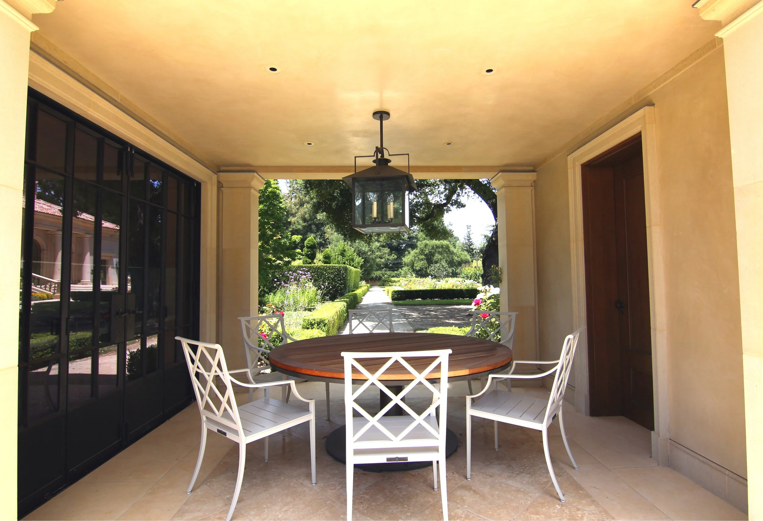 Outdoor covered patio with a round wooden table and six white chairs, hanging lantern light, and view of a garden with trees and bushes.