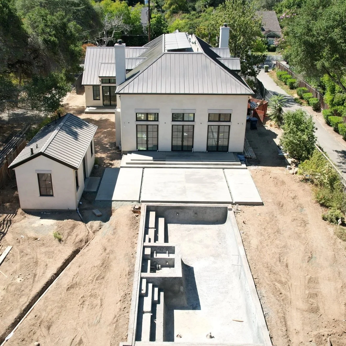 Aerial view of a residential construction site showing an unfinished backyard pool, a patio, a house with large windows, a small separate building, and dirt surroundings.
