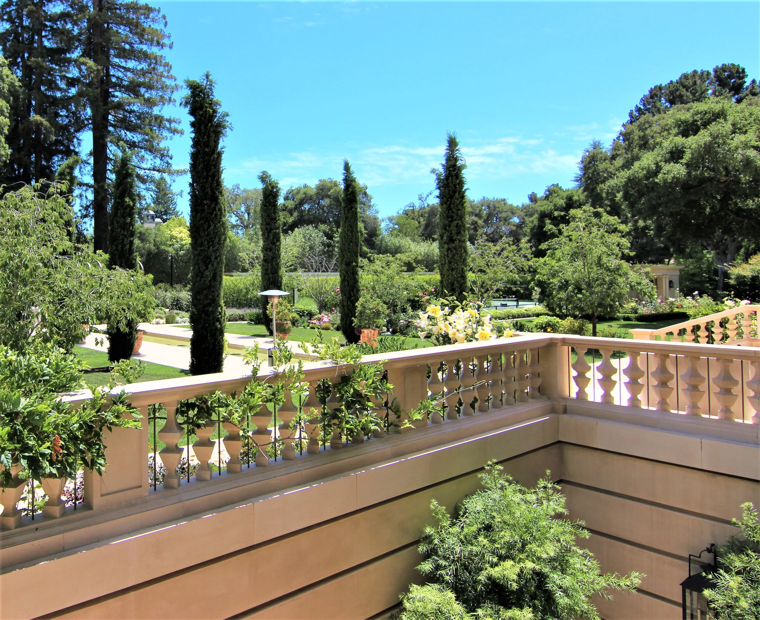 View from a balcony overlooking a lush, landscaped garden with tall cypress trees, various shrubs, and small trees, under a bright blue sky.