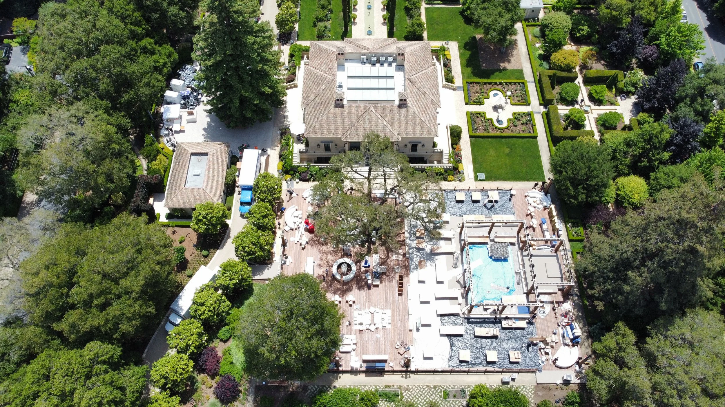 Aerial view of a large, luxurious mansion with a rooftop pool, outdoor seating area under a large tree, indoor construction in progress, surrounded by extensive manicured gardens, trees, hedges, and outdoor paths.