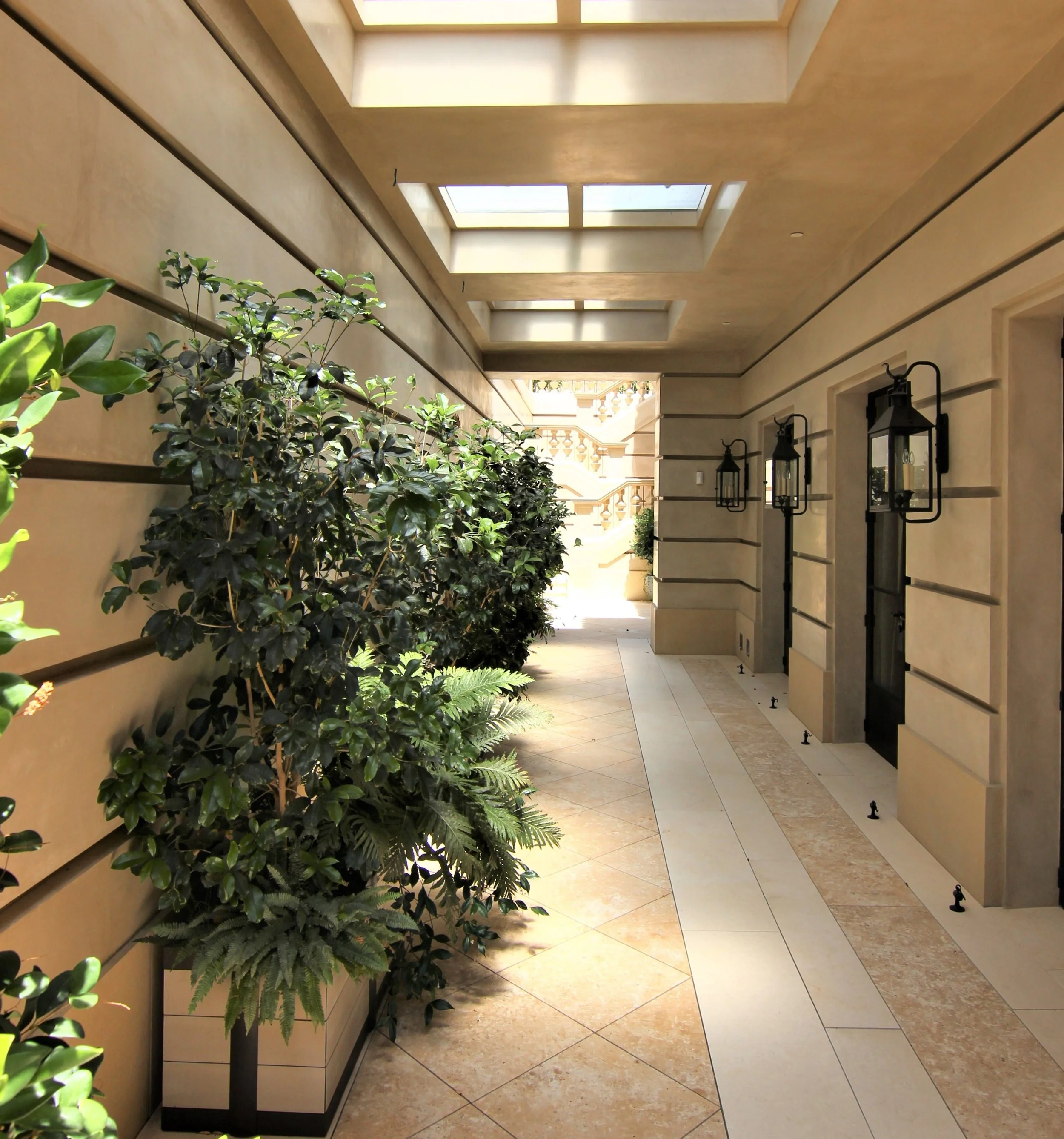 Covered outdoor walkway with potted plants on the left and black wall-mounted lanterns on the right wall, beige tile flooring, and rectangular skylights in the ceiling letting in natural sunlight.