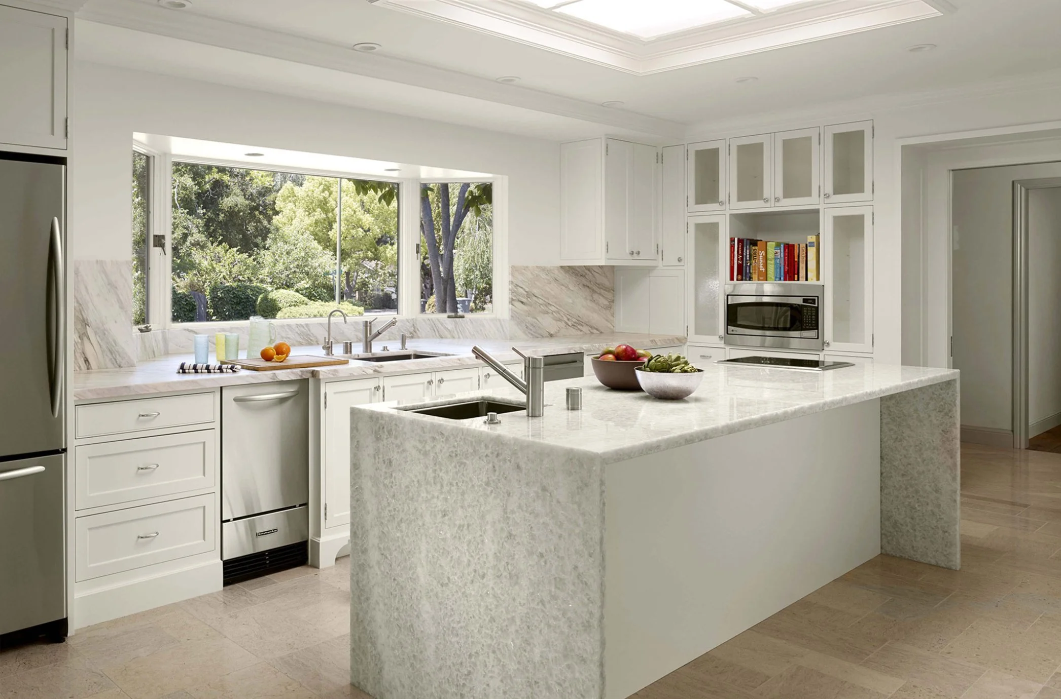 Modern kitchen with white cabinetry, marble countertops, and an island, large window showing greenery outside, with fruit bowls and kitchen appliances.