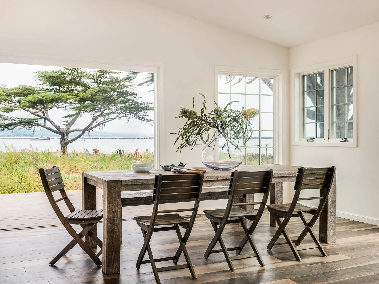 Interior of a dining room with a large wooden table, six wooden chairs, a glass vase with green plants, and a view of trees and water outside through large windows.