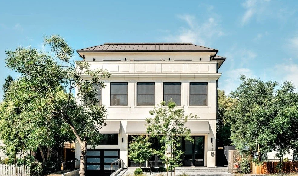 A three-story white house with large front windows and small trees in the front yard, under a partly cloudy blue sky.
