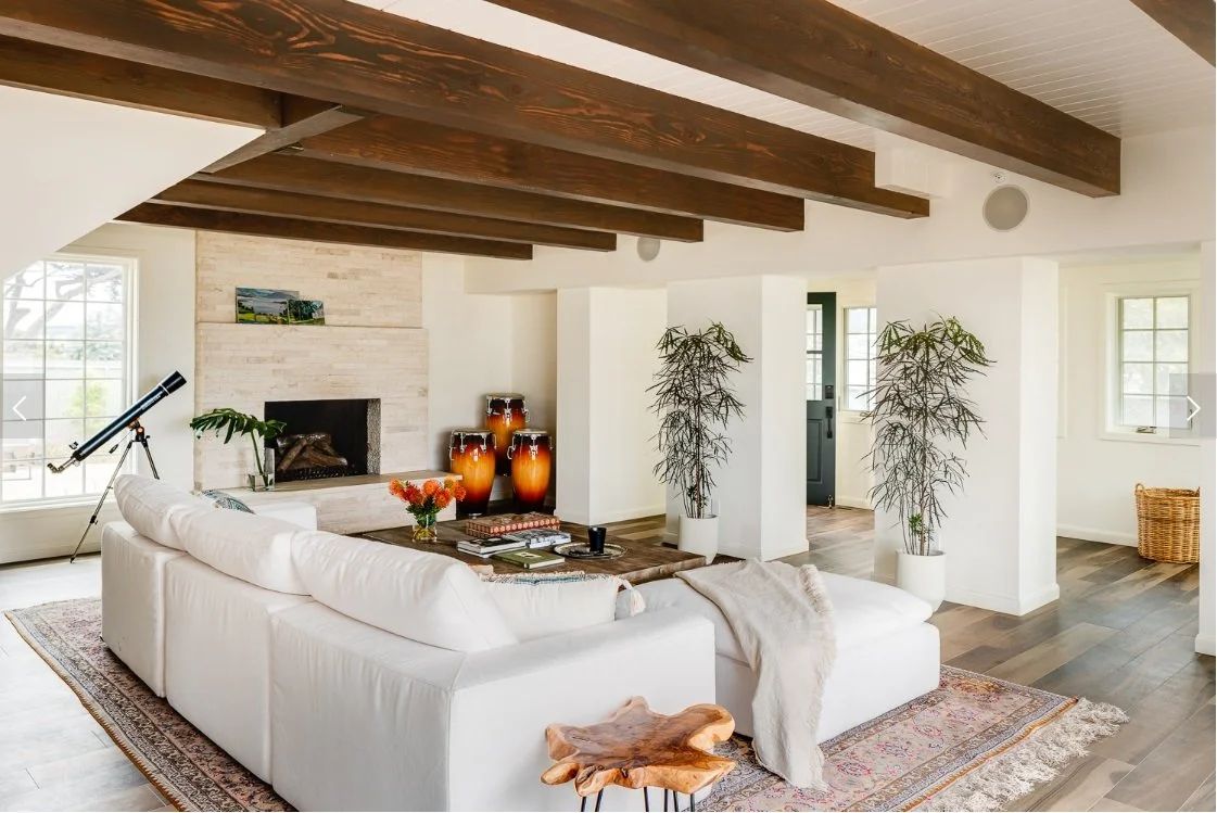 Bright living room with white walls and ceiling beams, large windows, white sofa, telescope, fireplace, potted plants, and decorative vases.
