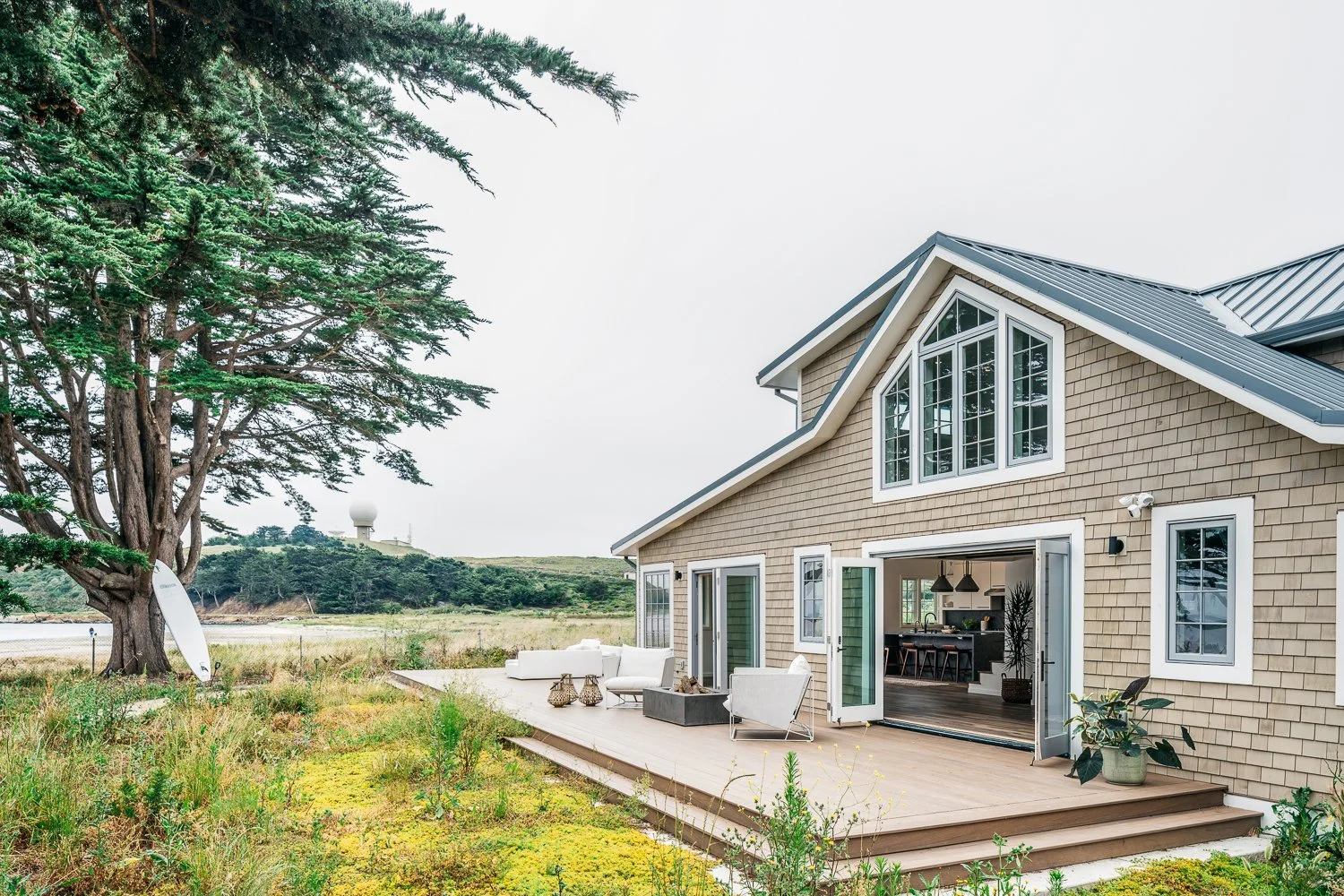 Exterior view of a modern house with a wooden deck surrounded by nature, a large tree on the left, and a distant hill topped with a radar station. The house has large windows and open sliding doors leading to the deck, which has outdoor furniture.