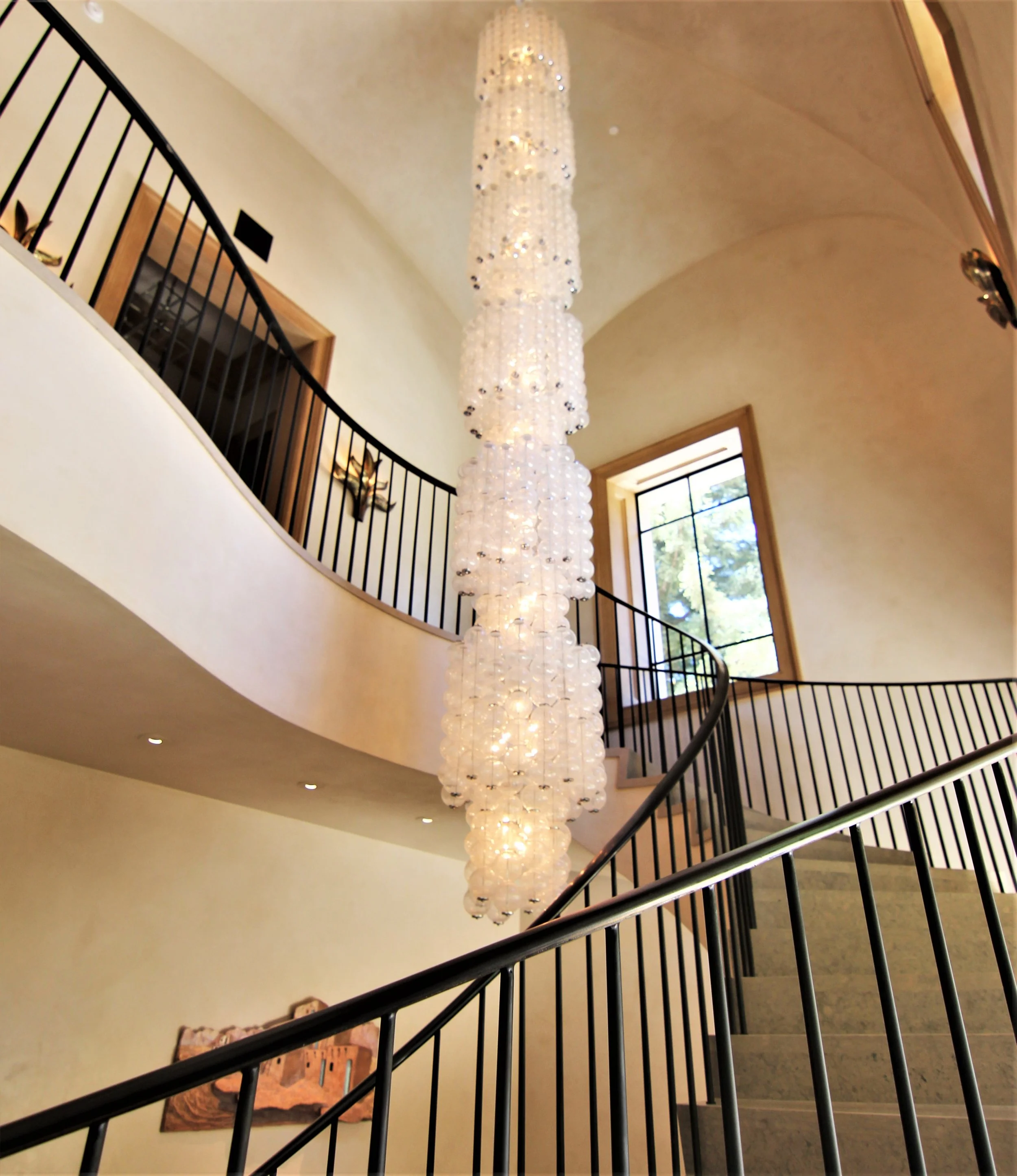 Elegant interior staircase with black metal railing, large chandelier composed of multiple glass or crystal globes, high ceiling, and a window letting in natural light.