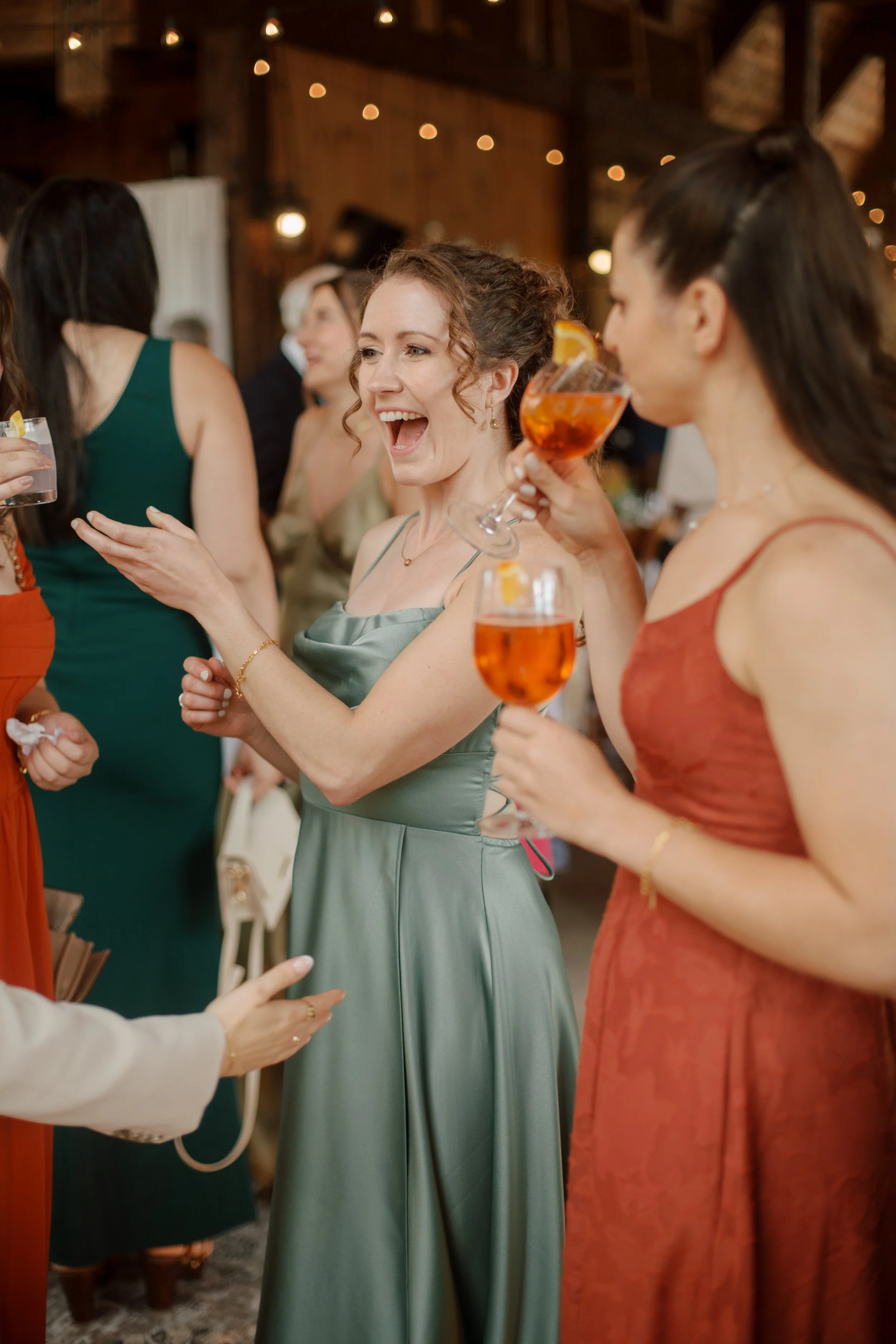 Women at a celebration holding drinks, smiling, and talking in a rustic indoor setting with warm lighting.
