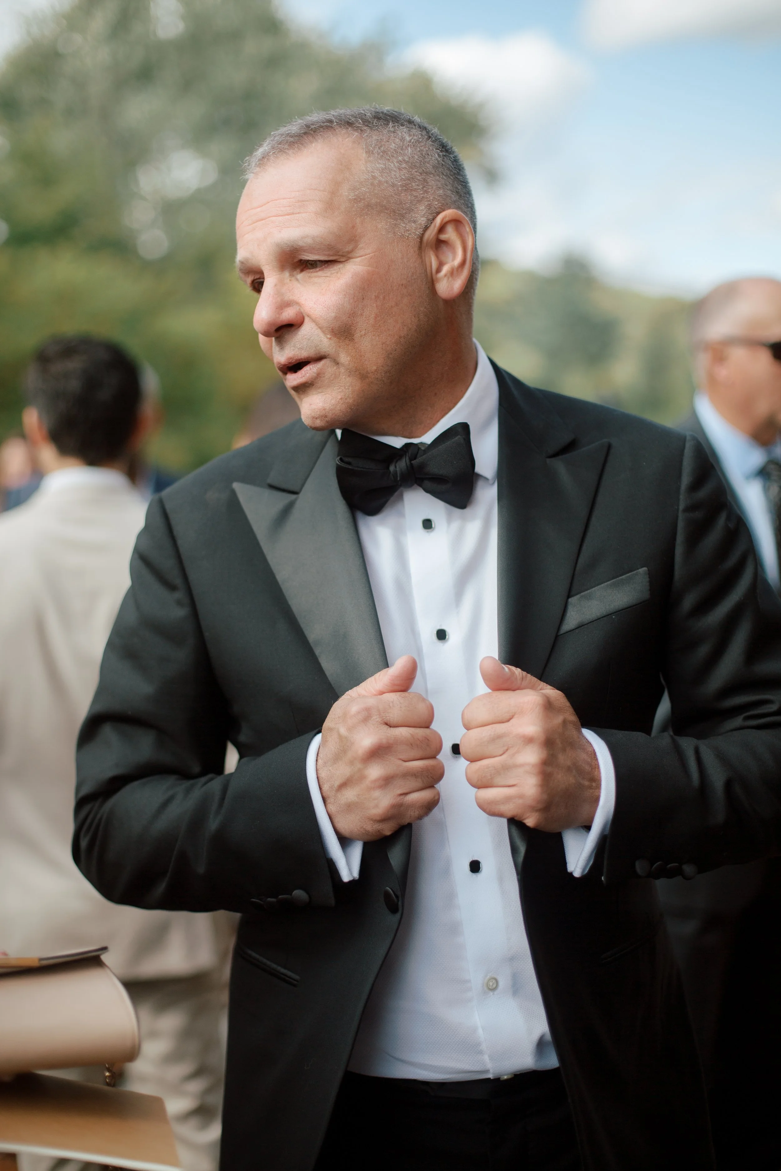A man in a tuxedo with a bowtie adjusting his jacket outdoors during the daytime, with people in the background.