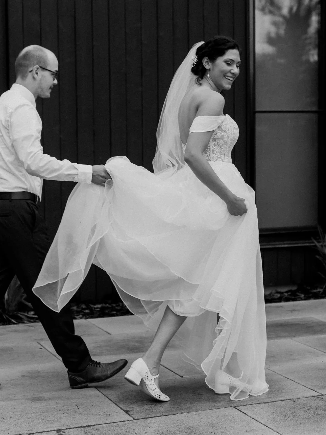 A bride in a wedding dress and veil smiling, being playfully pulled by a man in a white shirt and dark pants, outside in front of a black wooden wall.