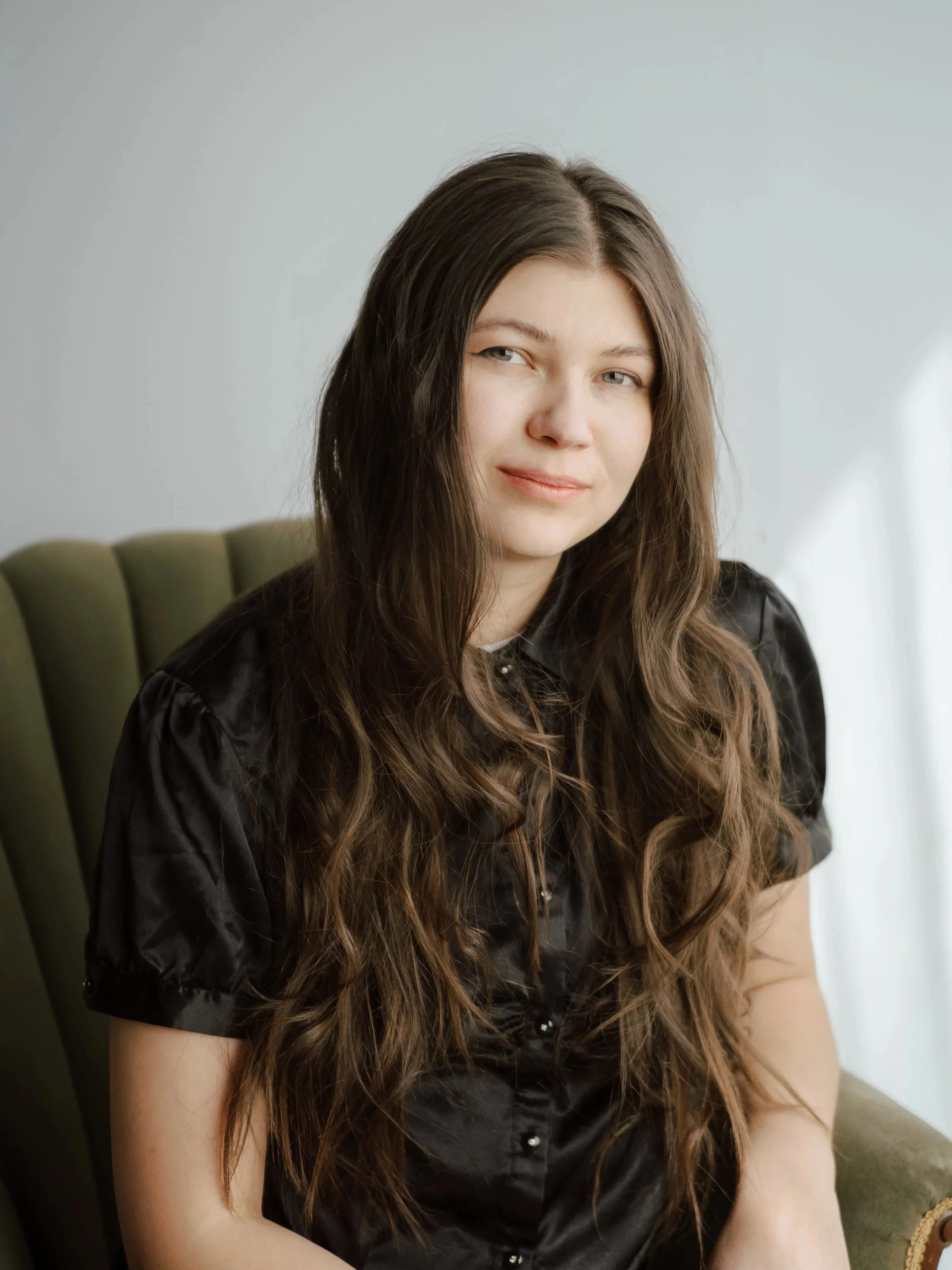 A young woman with long wavy hair, wearing a black shirt, sitting on a green chair against a plain light-colored wall.