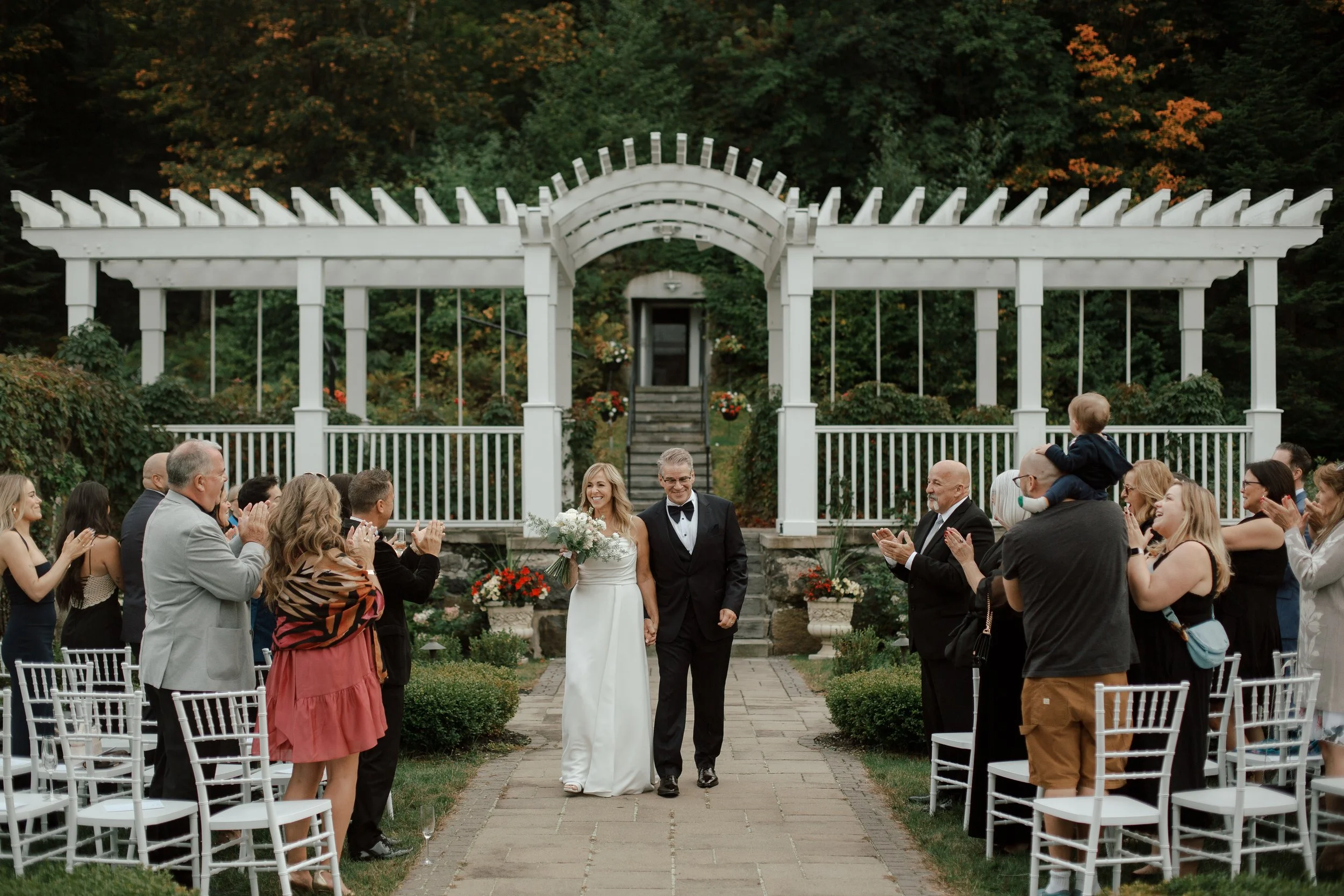 Bride and groom walking down the aisle at outdoor wedding ceremony, surrounded by seated guests clapping, set in a garden with white structure and green trees in background.