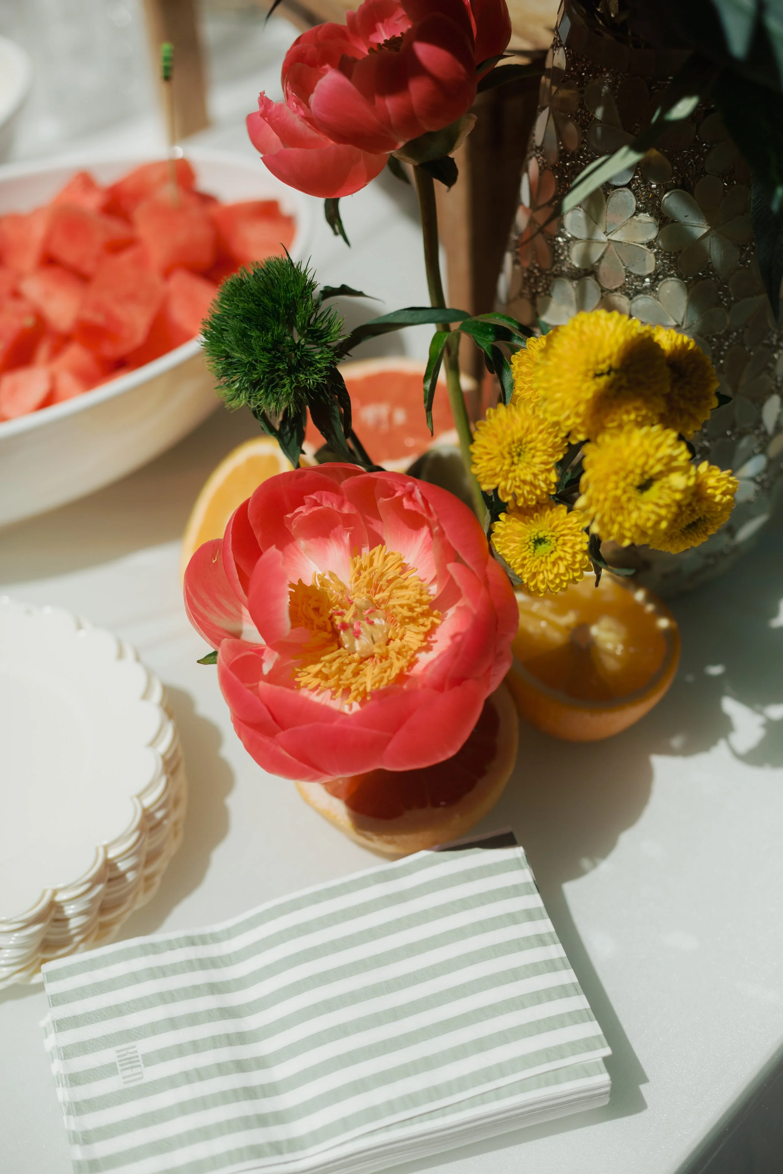 Close-up of a floral arrangement with pink peonies, yellow chrysanthemums, and green foliage in a decorative vase, with slices of grapefruit and a bowl of watermelon in the background, and a stack of white and pale green striped napkins in the foreground.