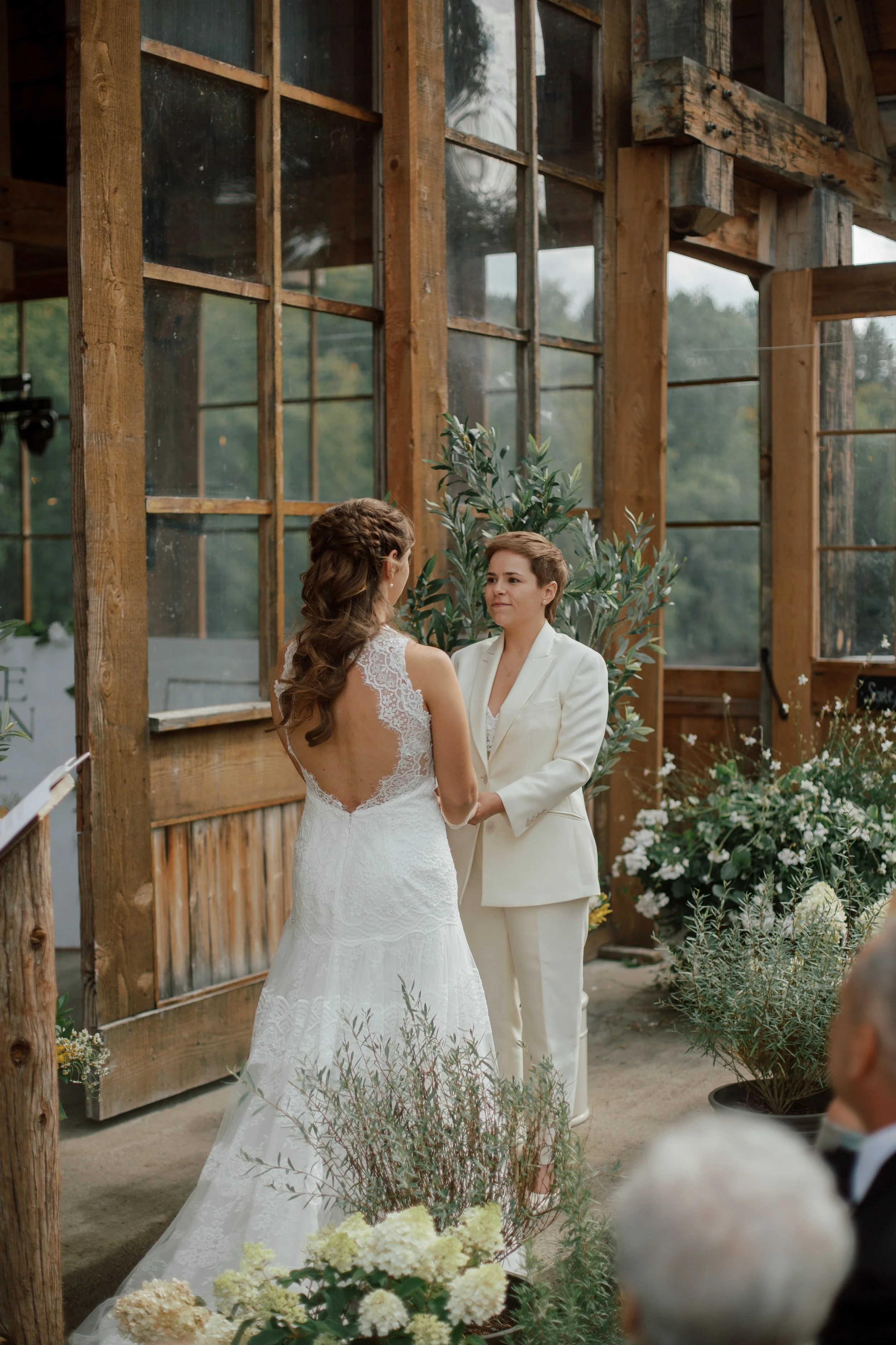 Two women holding hands during a wedding ceremony, one in a white lace wedding dress and the other in a white pantsuit, inside a rustic wooden and glass structure decorated with plants and flowers.