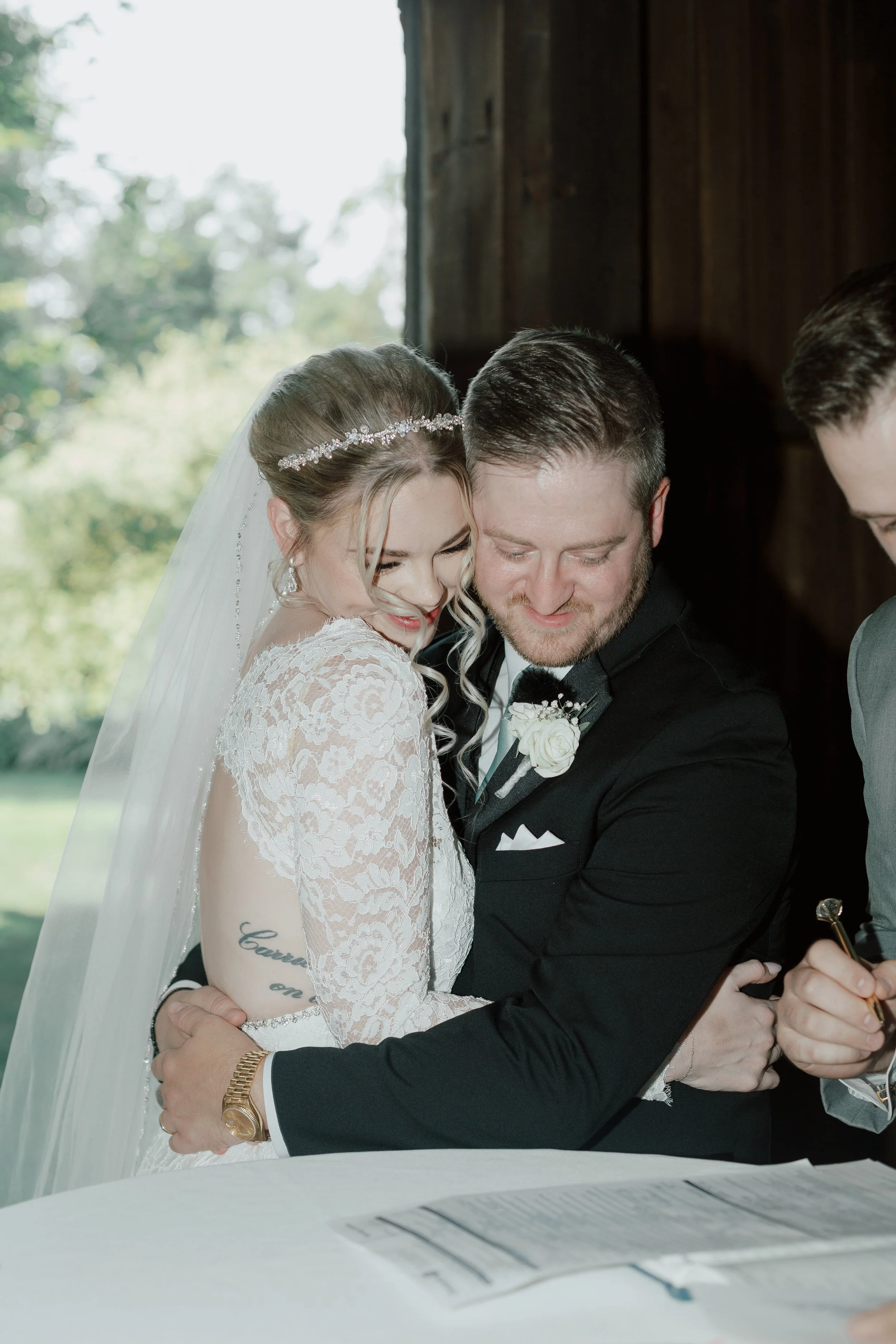 A bride and groom are smiling and holding hands as they sign their marriage certificate, with a person partially visible on the right side of the image.