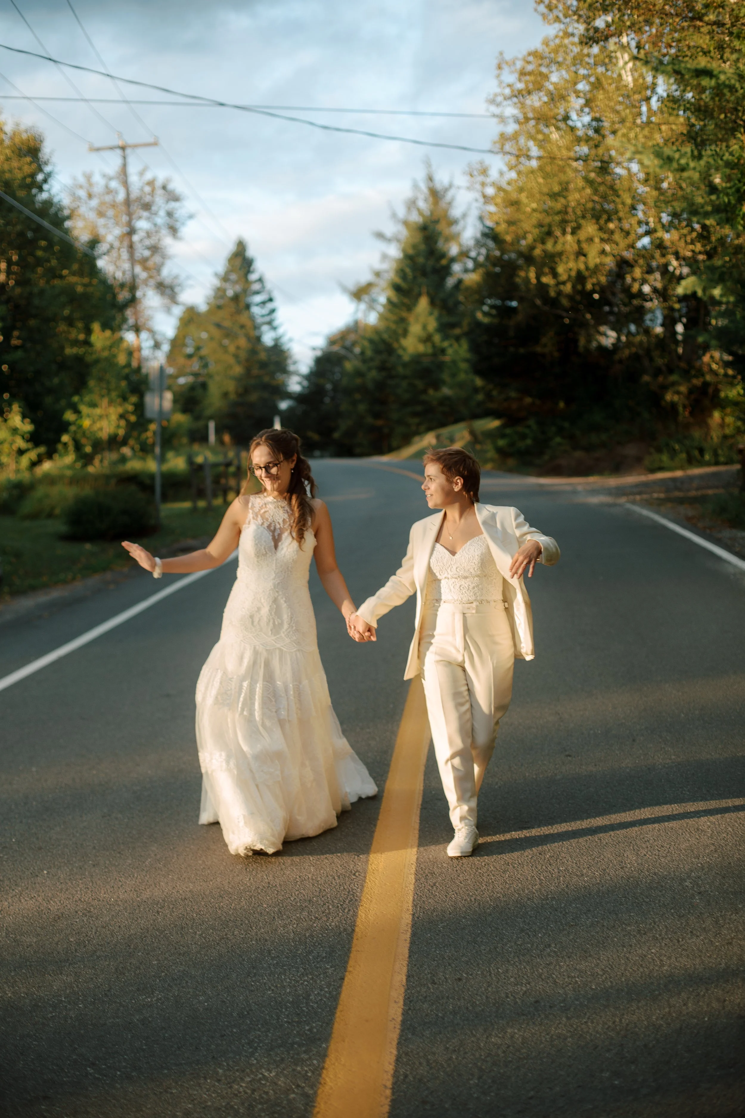 Two women, one in a wedding dress and the other in a cream pantsuit, holding hands and walking on a road surrounded by trees during sunset.