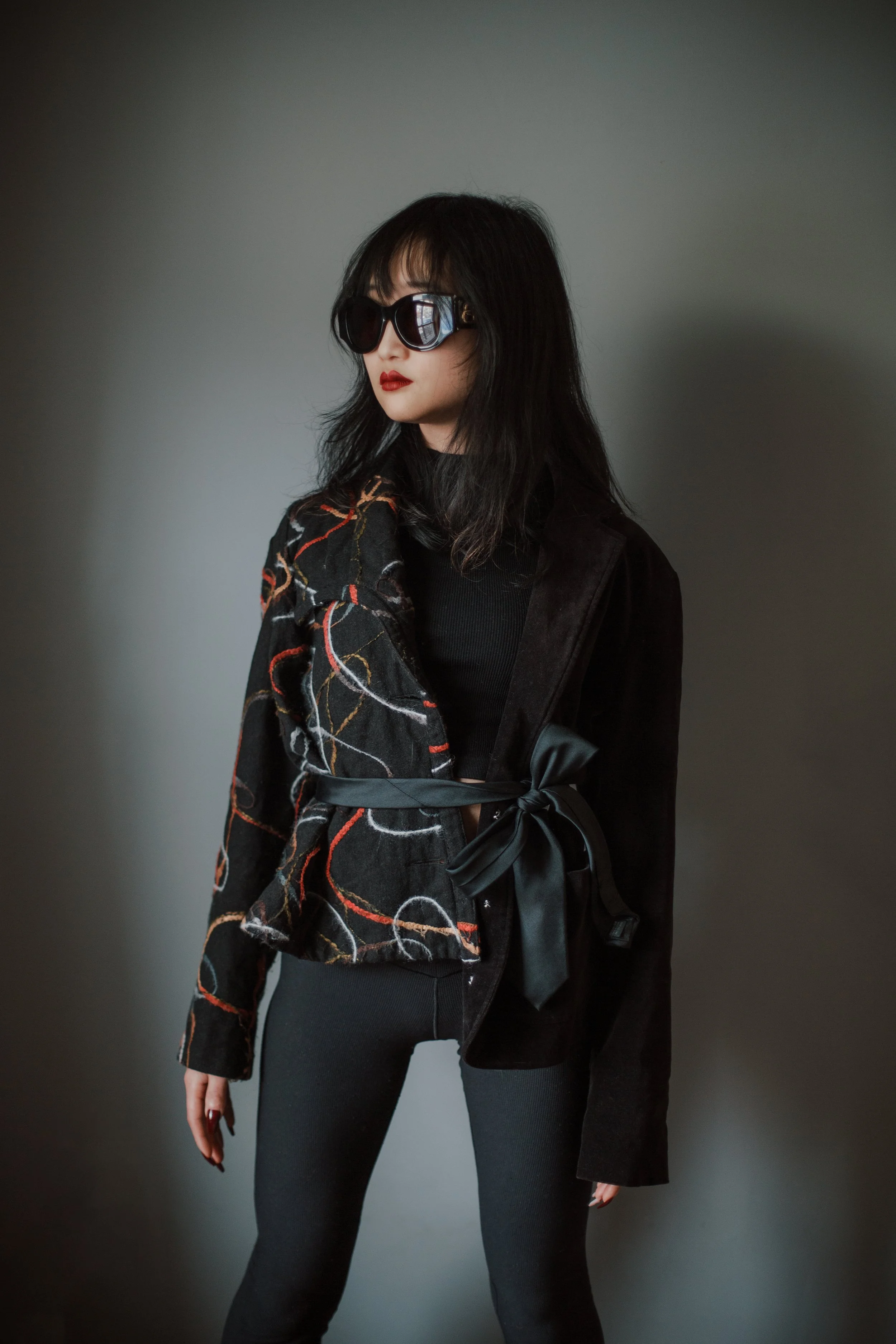 A woman with dark hair wearing large sunglasses, a black top, and a black jacket tied with a satin ribbon in front, standing against a neutral gray background.