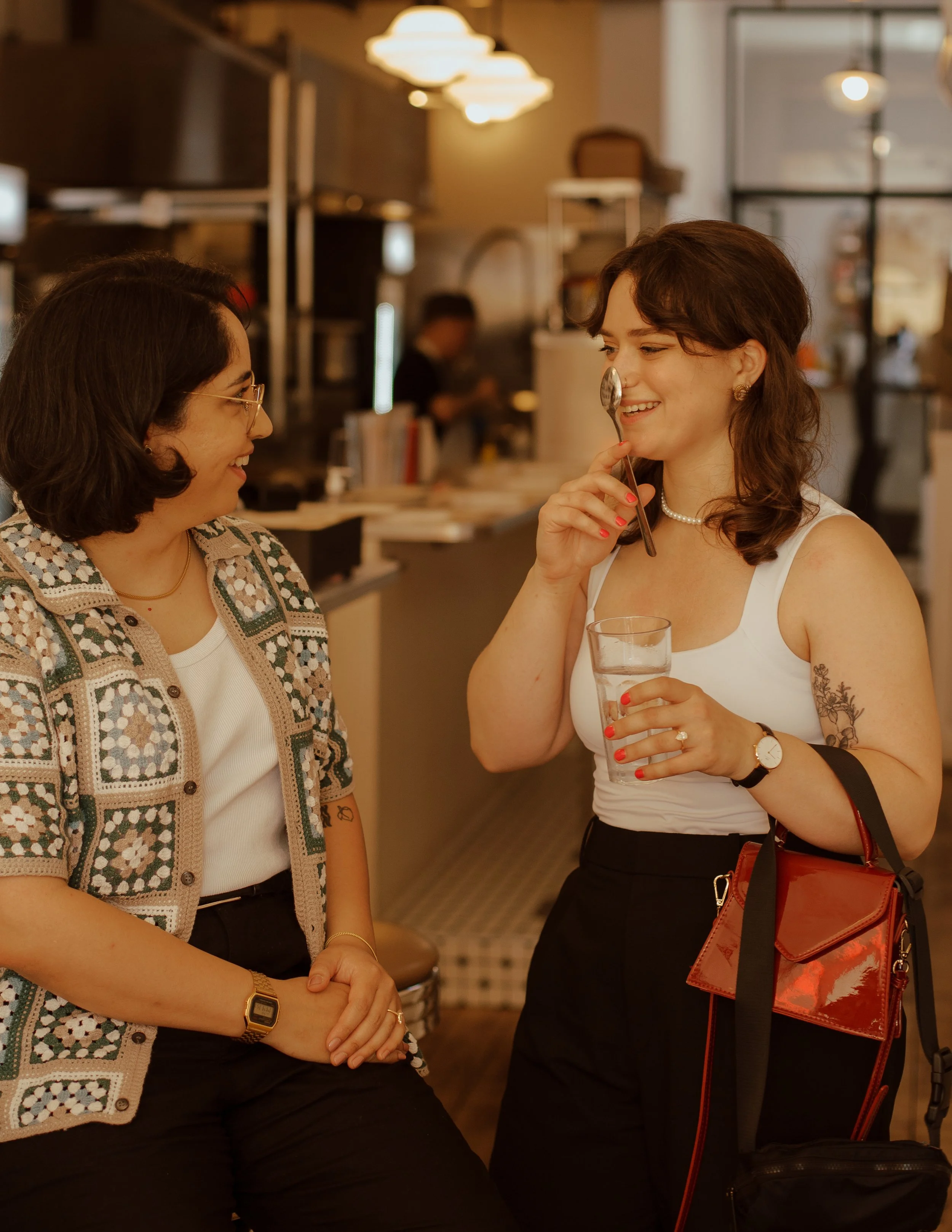 Two women talking and smiling in a café. One holds a glass of water, the other holds a spoon near her face. The background shows a barista and café equipment.