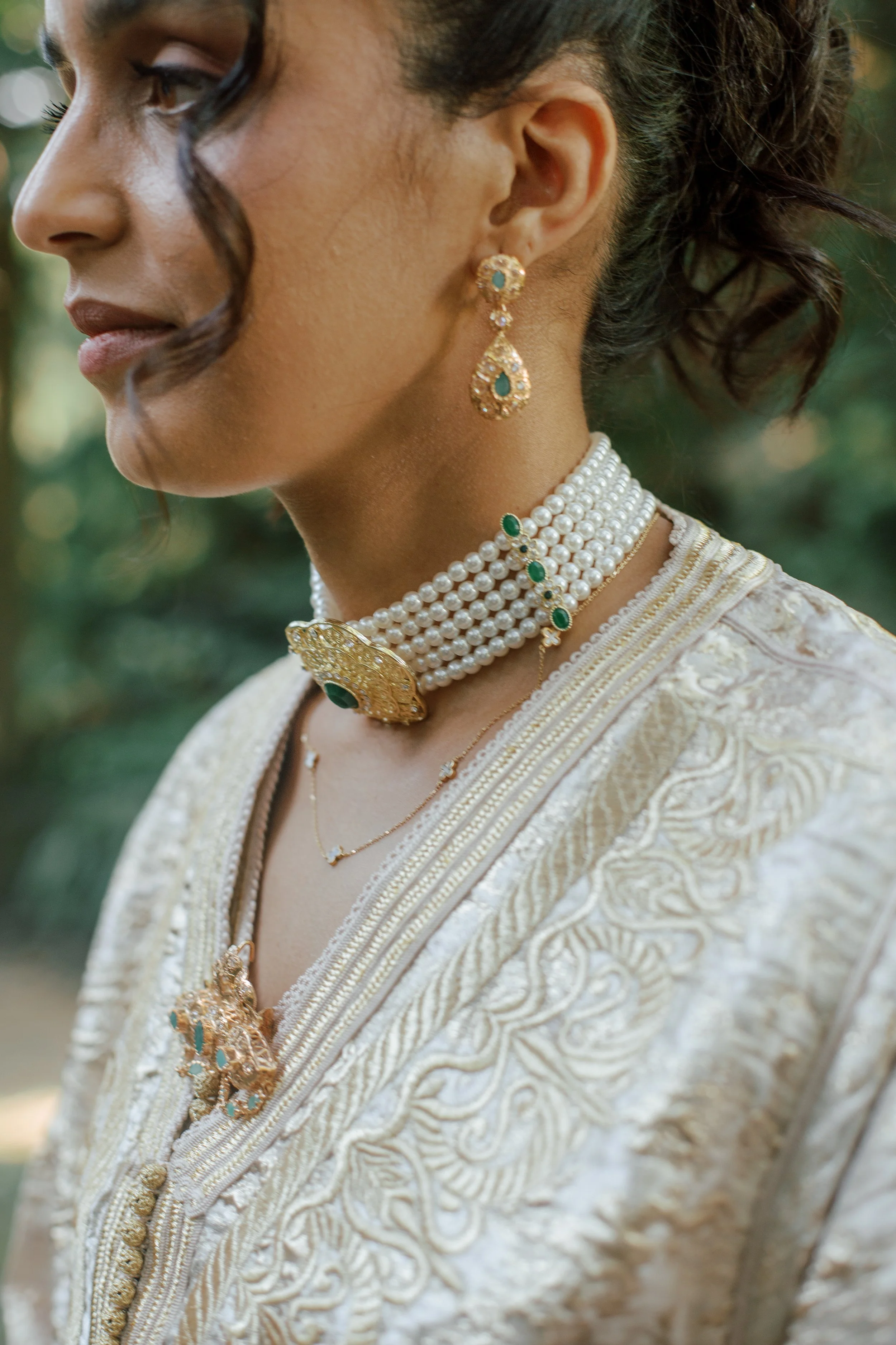 Close-up of a woman wearing traditional Indian jewelry and attire, including earrings, layered pearl necklaces, and a richly embroidered cream-colored dress.