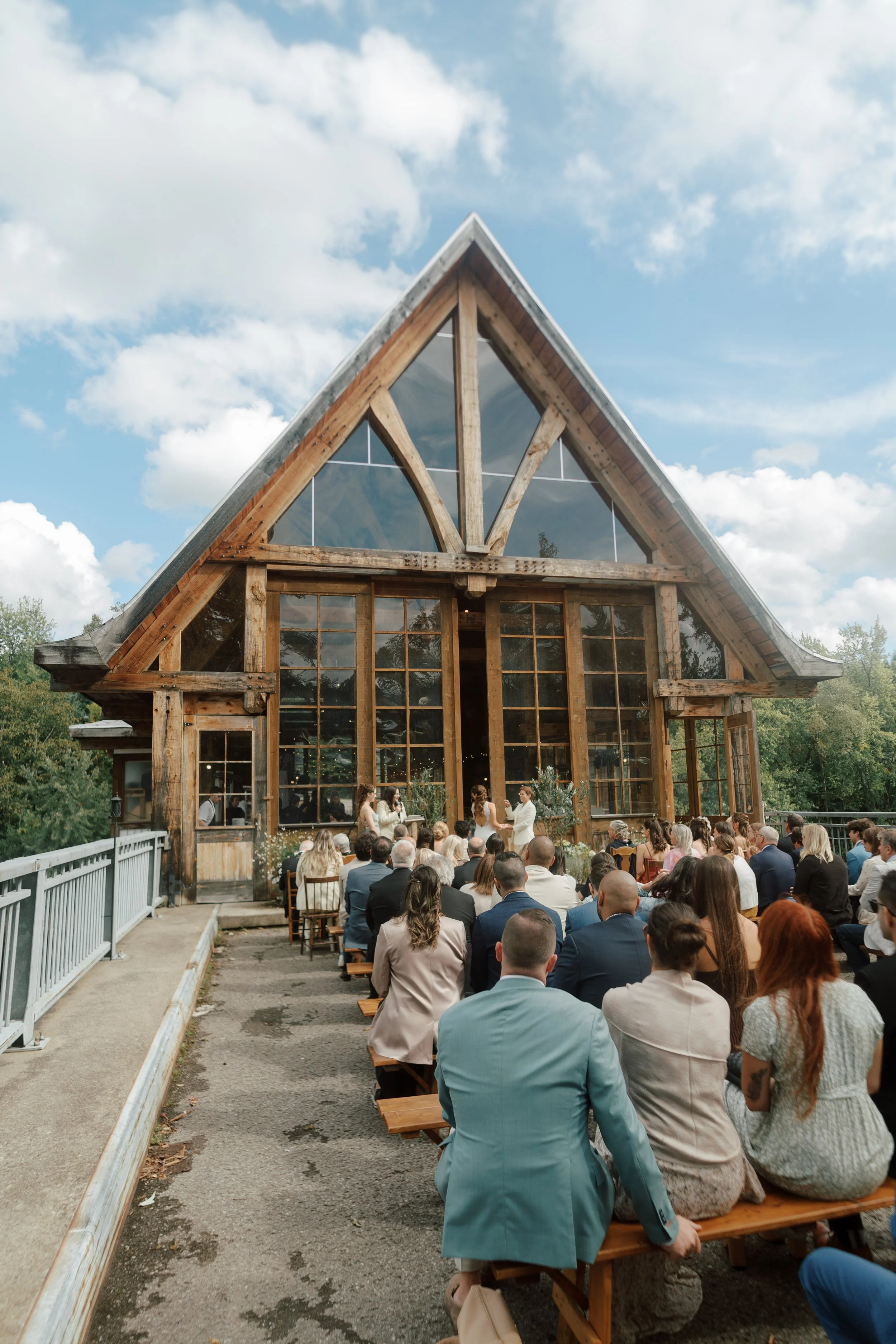 People attending a wedding ceremony outdoors in front of a large wooden A-frame building with glass windows. The sky is partly cloudy.