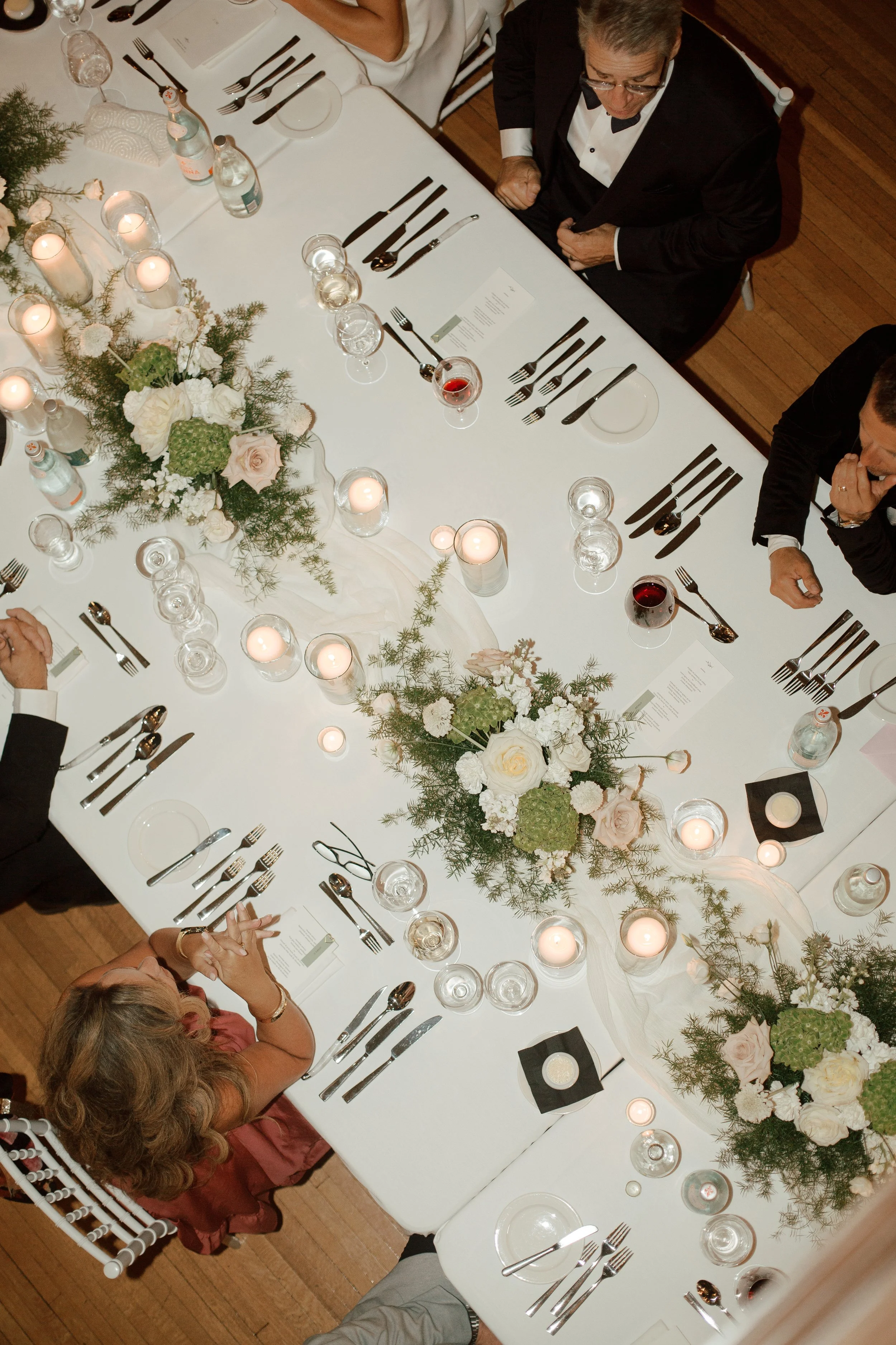 Overhead view of an elegant dinner table set with floral centerpieces, candles, wine glasses, and formal place settings, with guests seated in tuxedos and formal attire.