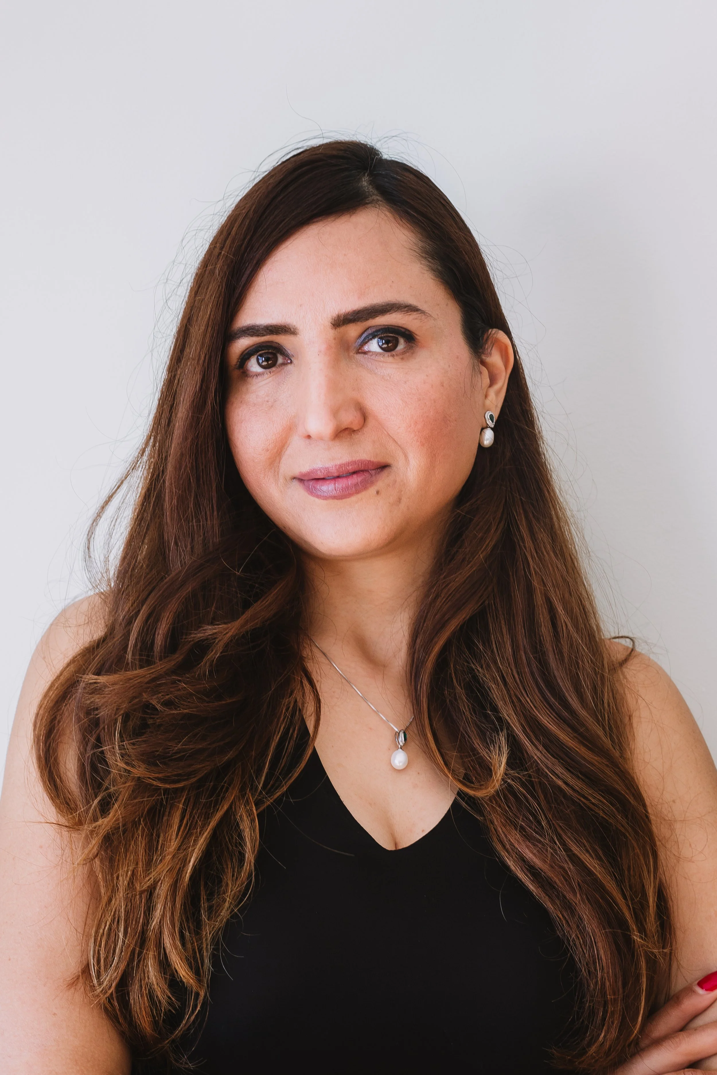 A woman with long, wavy brown hair wearing a black top, pearl jewelry, and a necklace with a pearl pendant, standing against a white background.