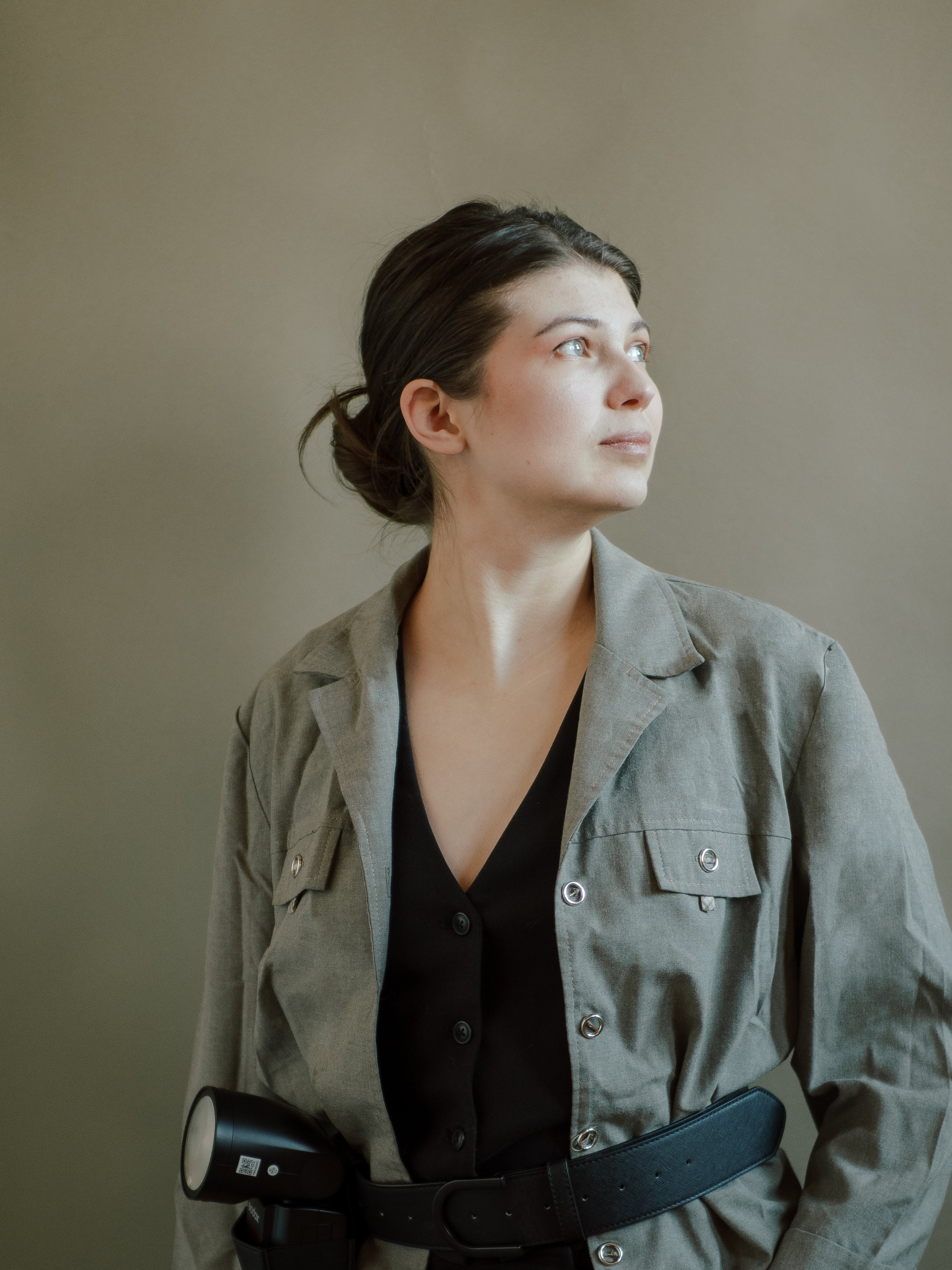 A woman with dark hair pulled back, wearing a gray jacket over a black top, standing against a plain wall, looking off to the side.