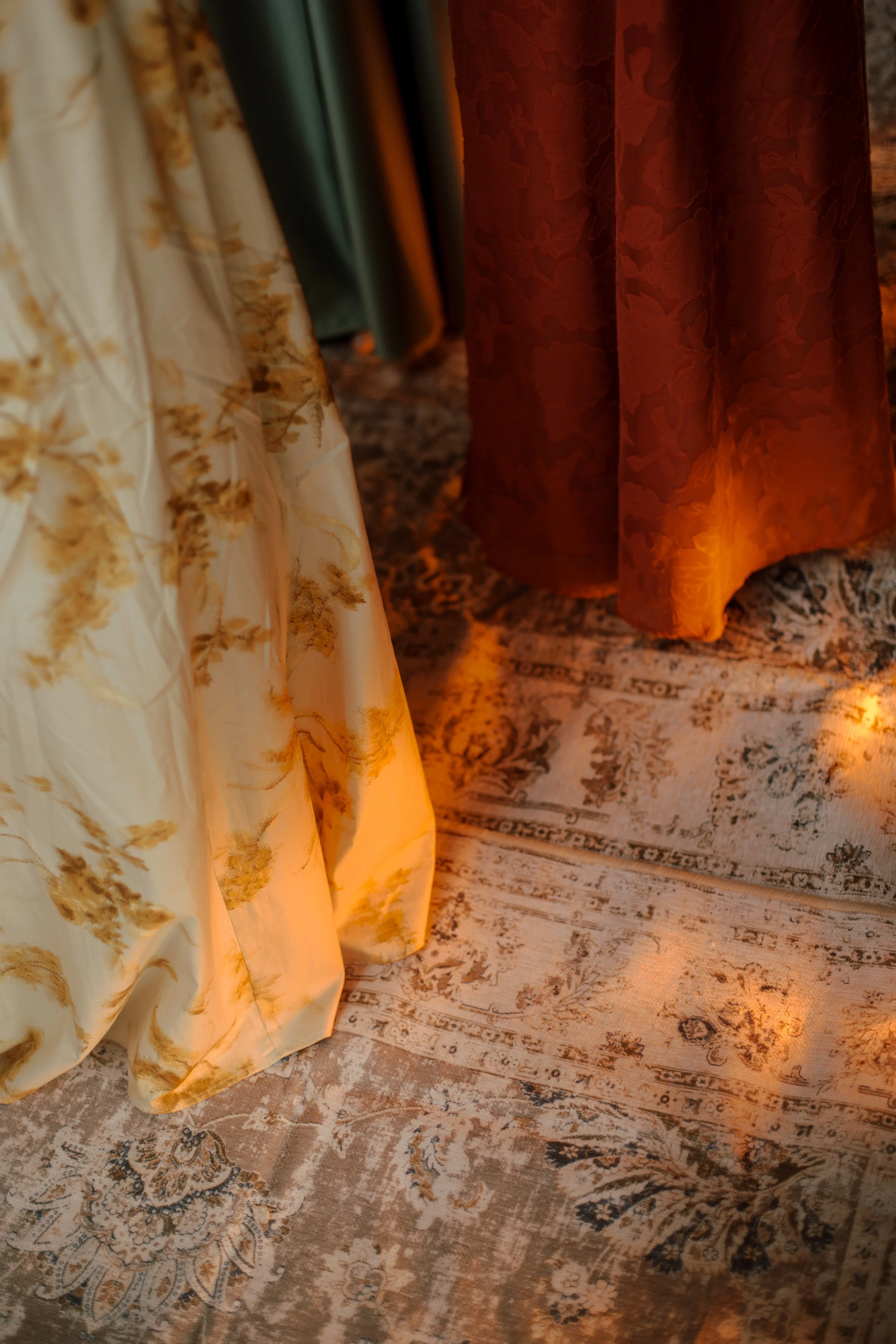 Close-up of several draped curtains in warm light, with a patterned rug underneath.