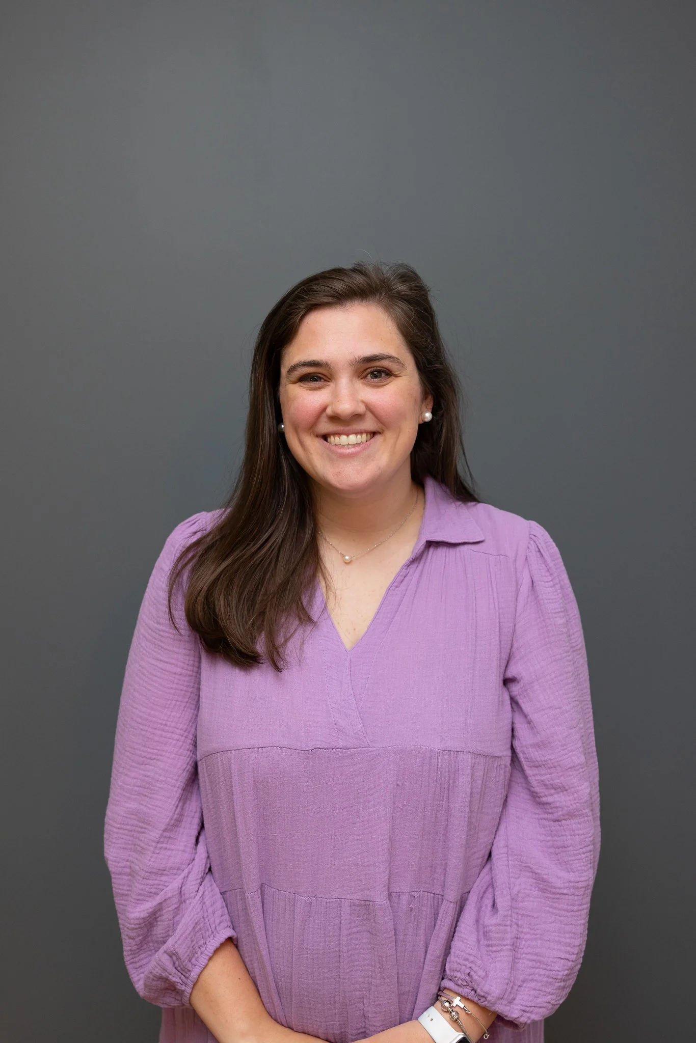 A woman with long brown hair smiling, wearing a light purple top, pearl earrings, a necklace, and bracelets, standing in front of a plain gray background.
