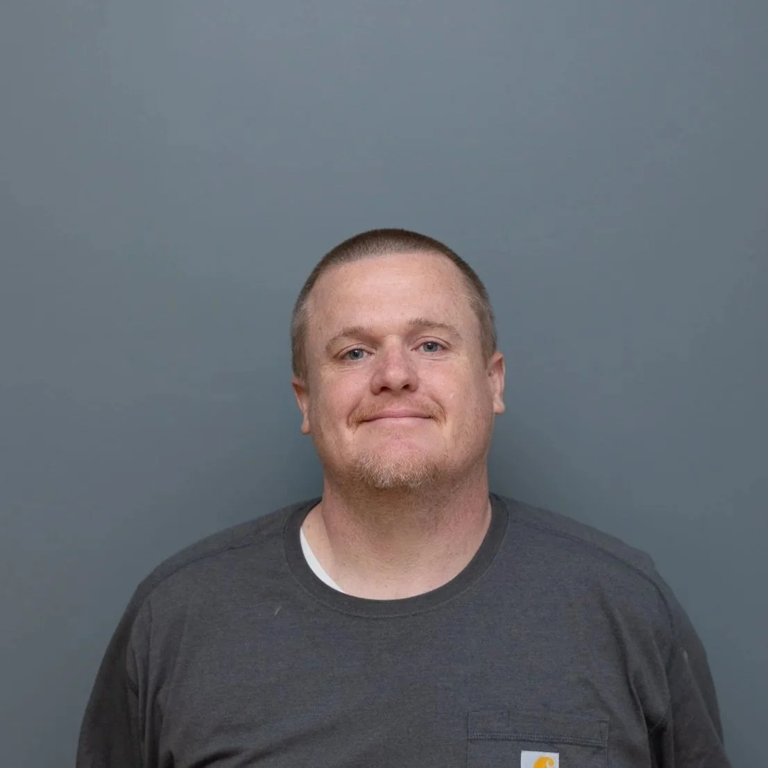 A young man with short light brown hair, a light beard, and blue eyes, smiling slightly, wearing a gray Carhartt t-shirt, standing against a plain blue-gray background.
