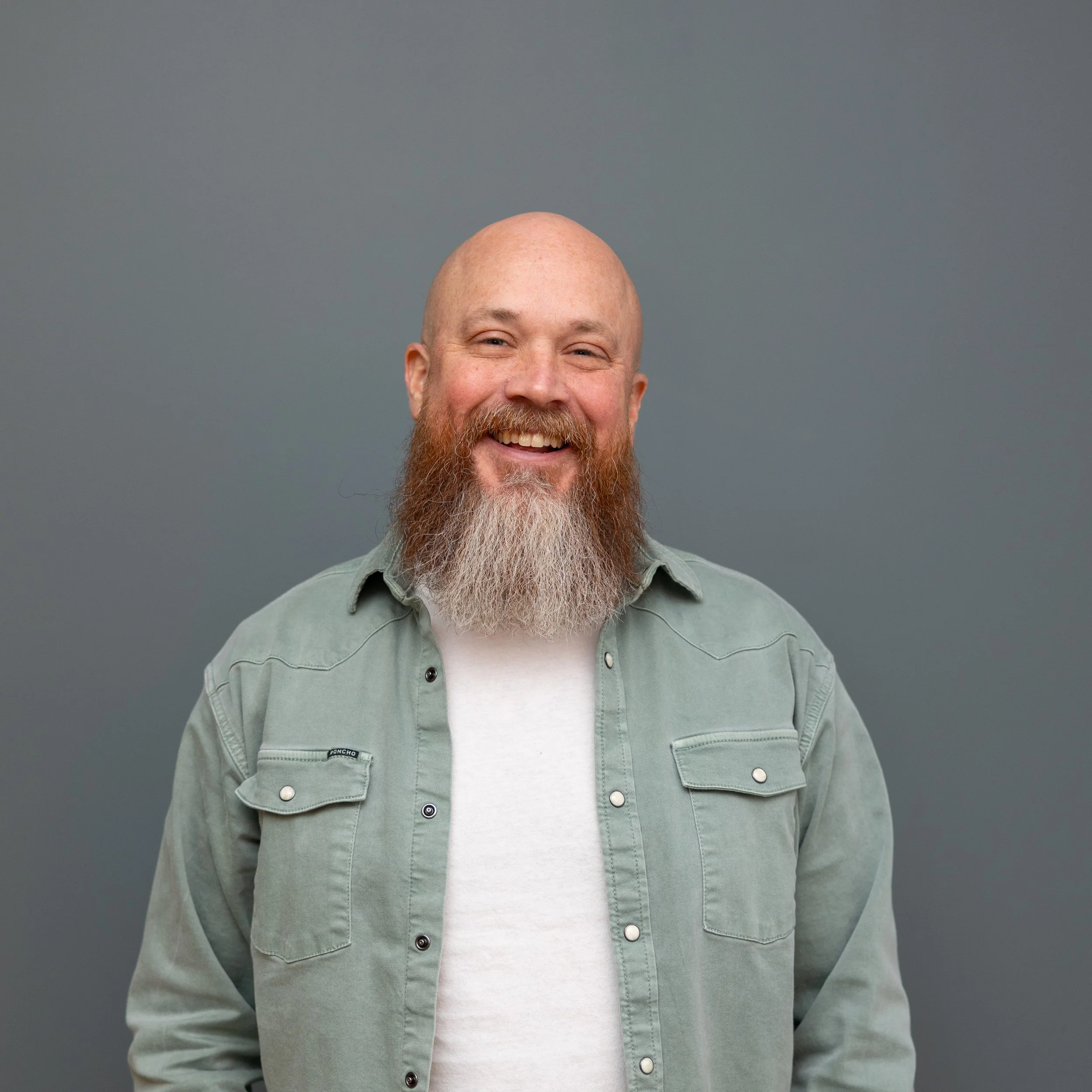 A smiling man with a bald head, reddish beard, and mustache, wearing a light green button-up shirt over a white T-shirt, standing against a plain gray background.
