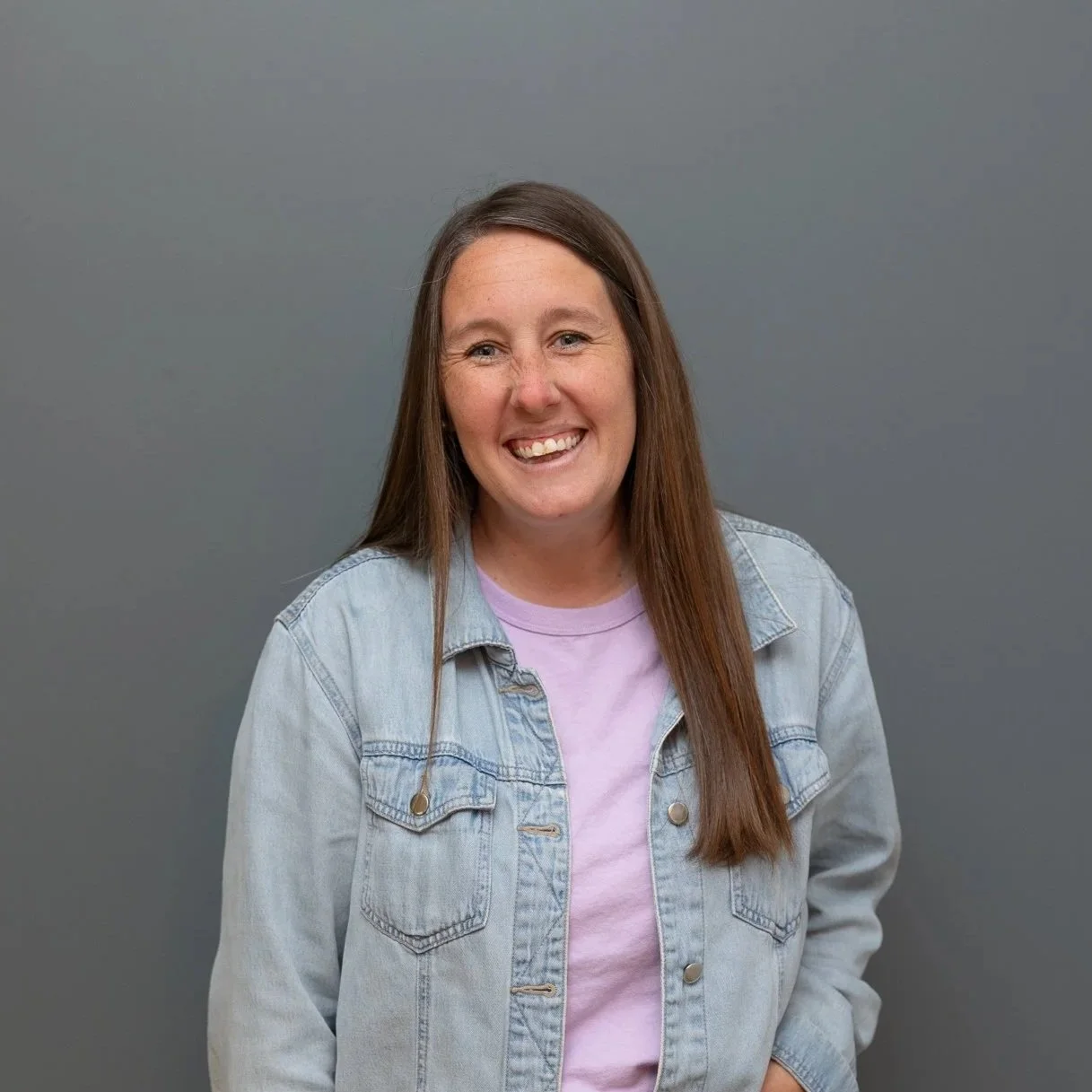 A smiling woman with long brown hair wearing a light denim jacket and a lavender T-shirt, standing against a neutral gray background.