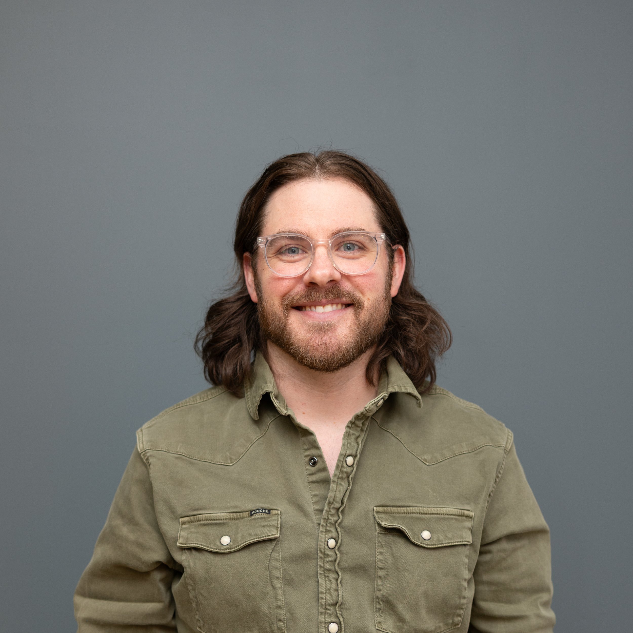 A portrait of a smiling man with shoulder-length brown hair, beard, and glasses, wearing a green button-up shirt, standing against a gray background.