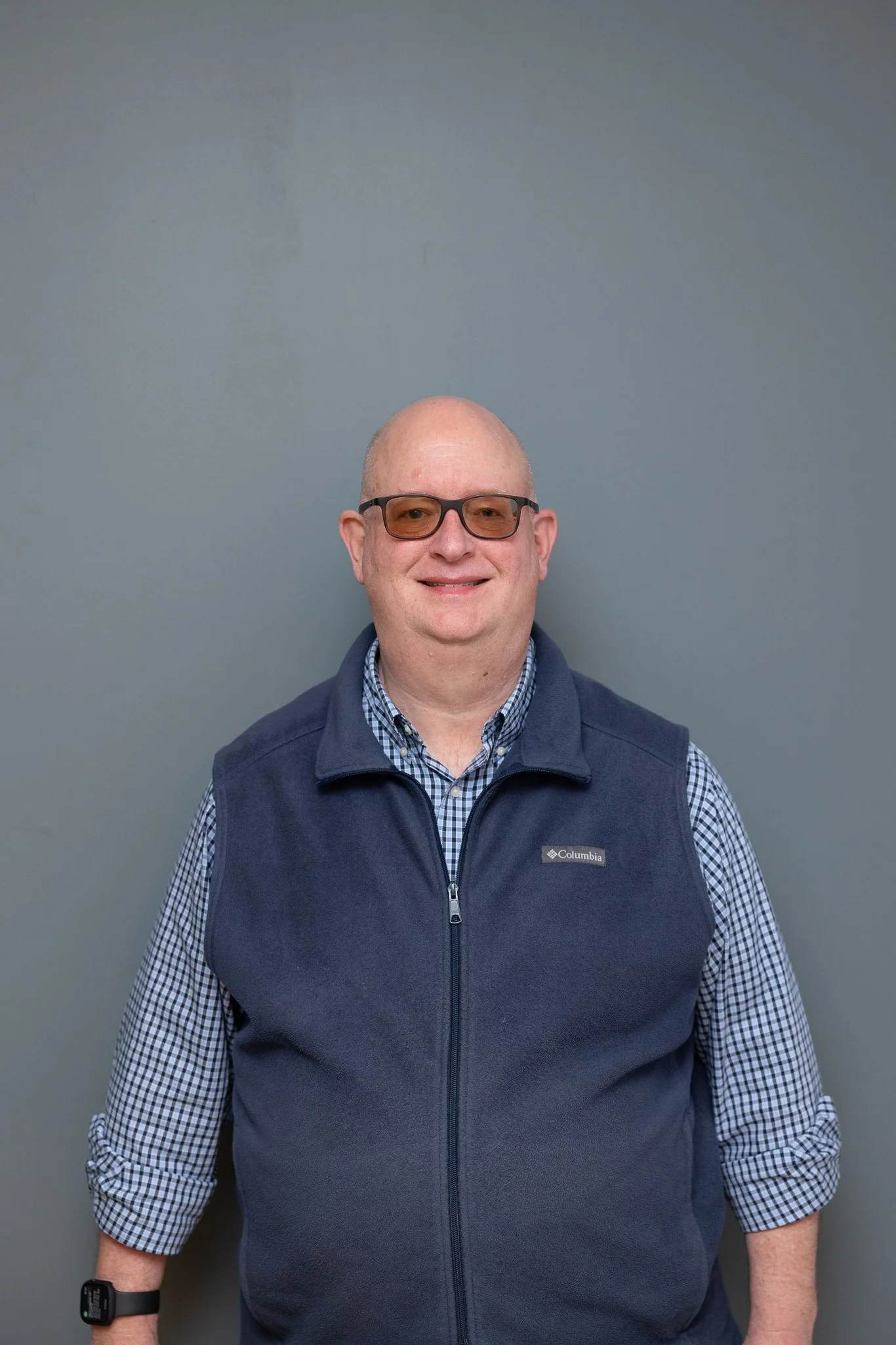 A man with glasses, wearing a checkered shirt and a navy blue Columbia vest, standing in front of a plain gray wall.