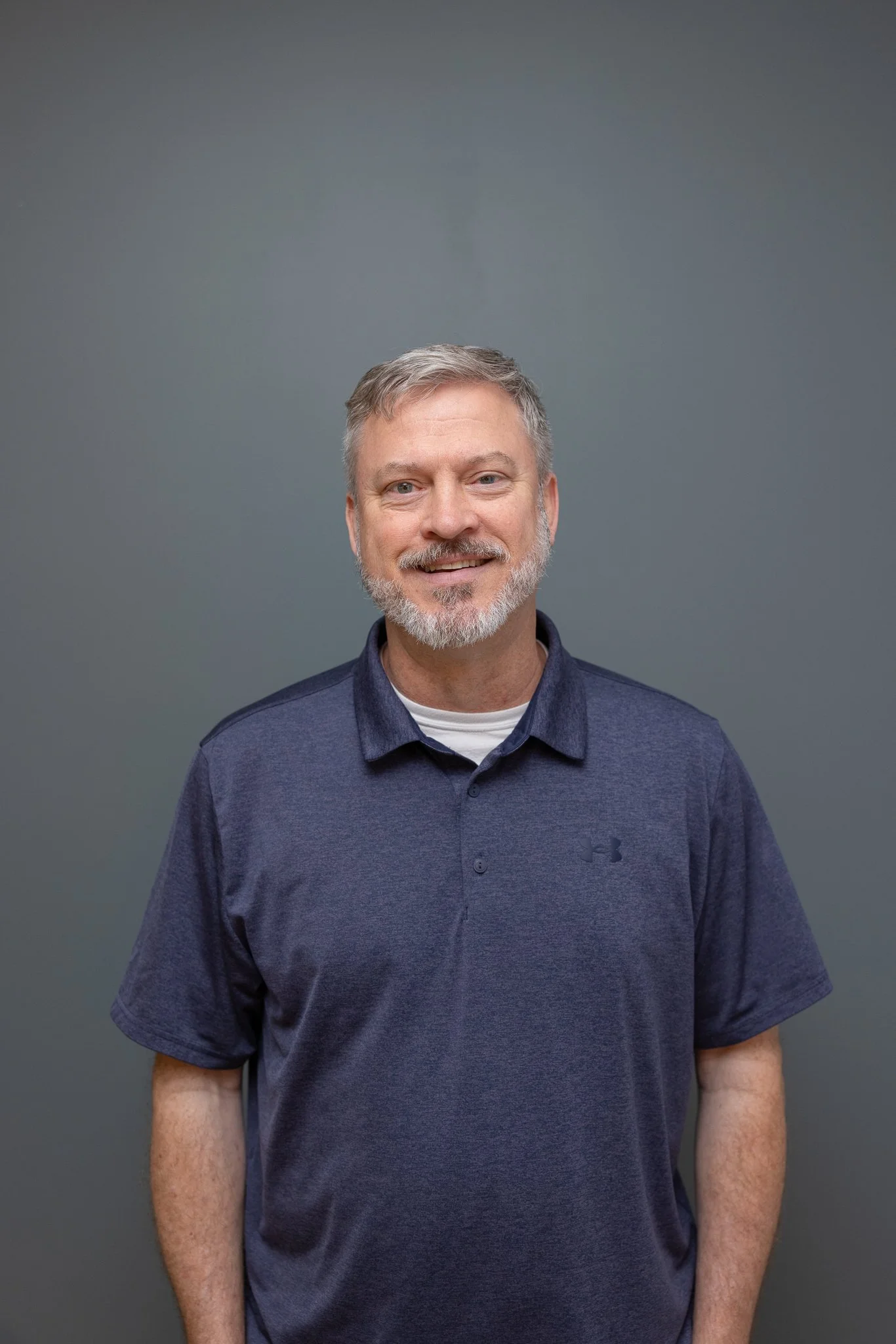 A middle-aged man with gray hair and a beard, smiling, wearing a navy blue collared Under Armour shirt, standing against a plain gray background.
