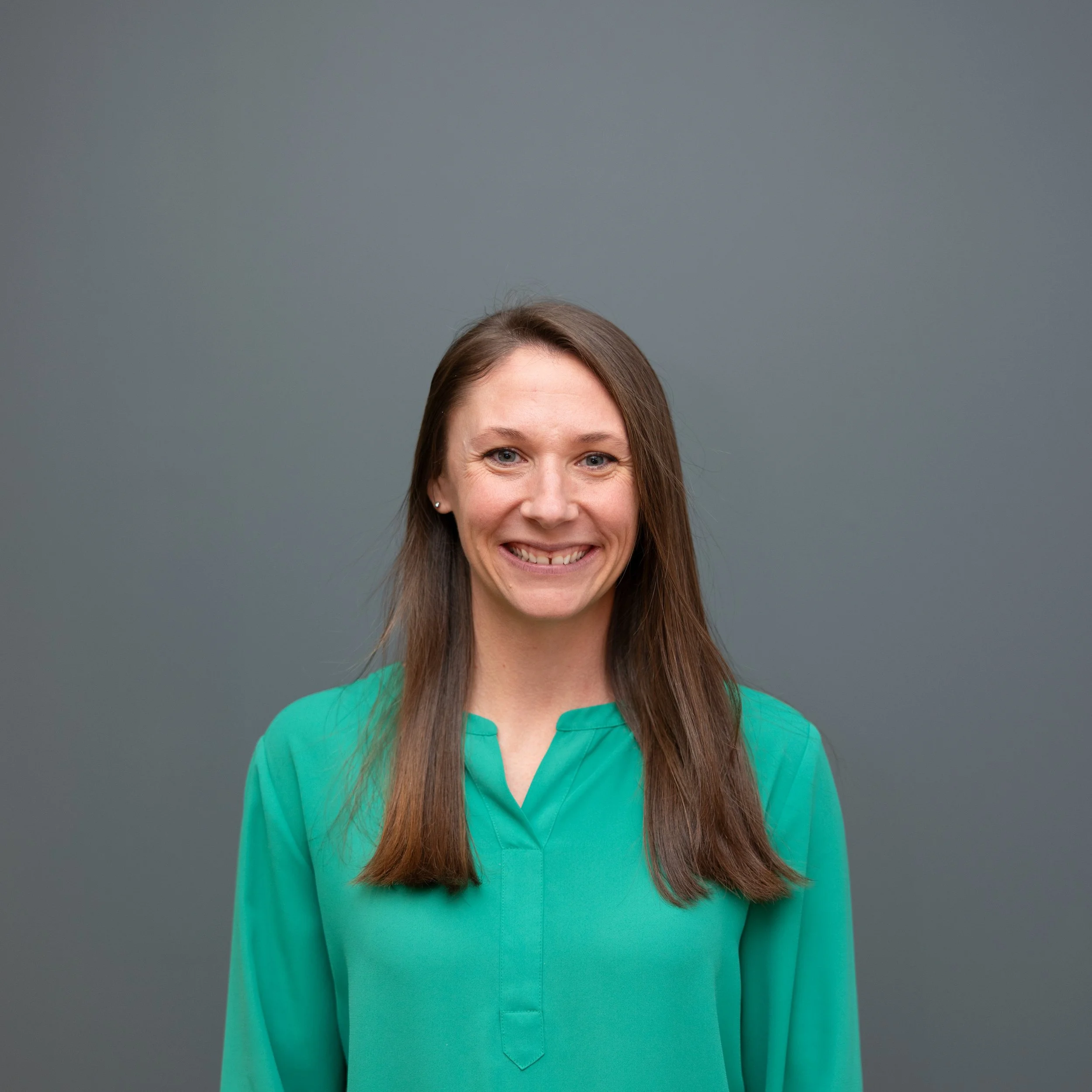 A woman with long brown hair smiling at the camera, wearing a green blouse, standing against a plain gray background.