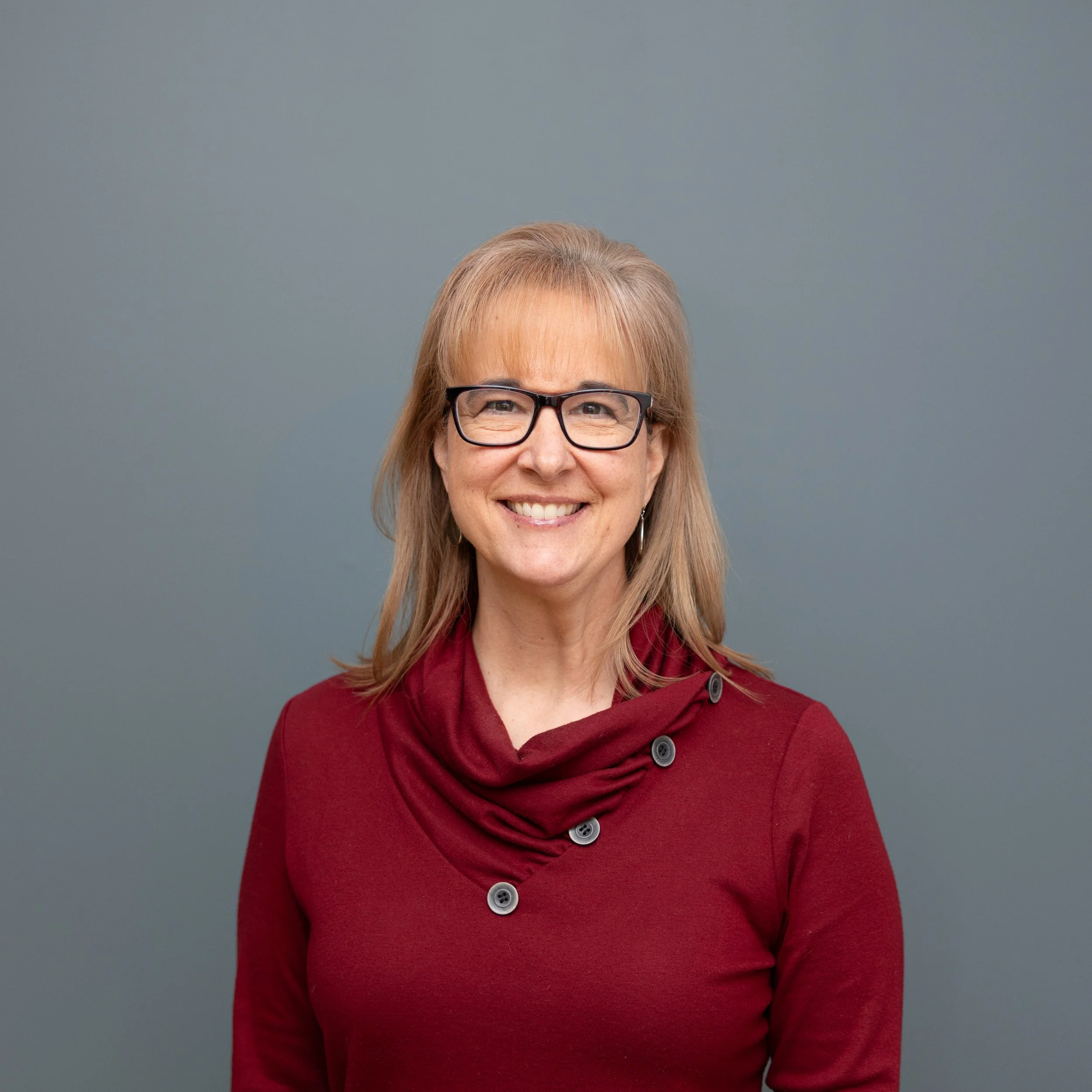 Portrait of a smiling woman with glasses, wearing a maroon top with buttons, standing against a gray background.